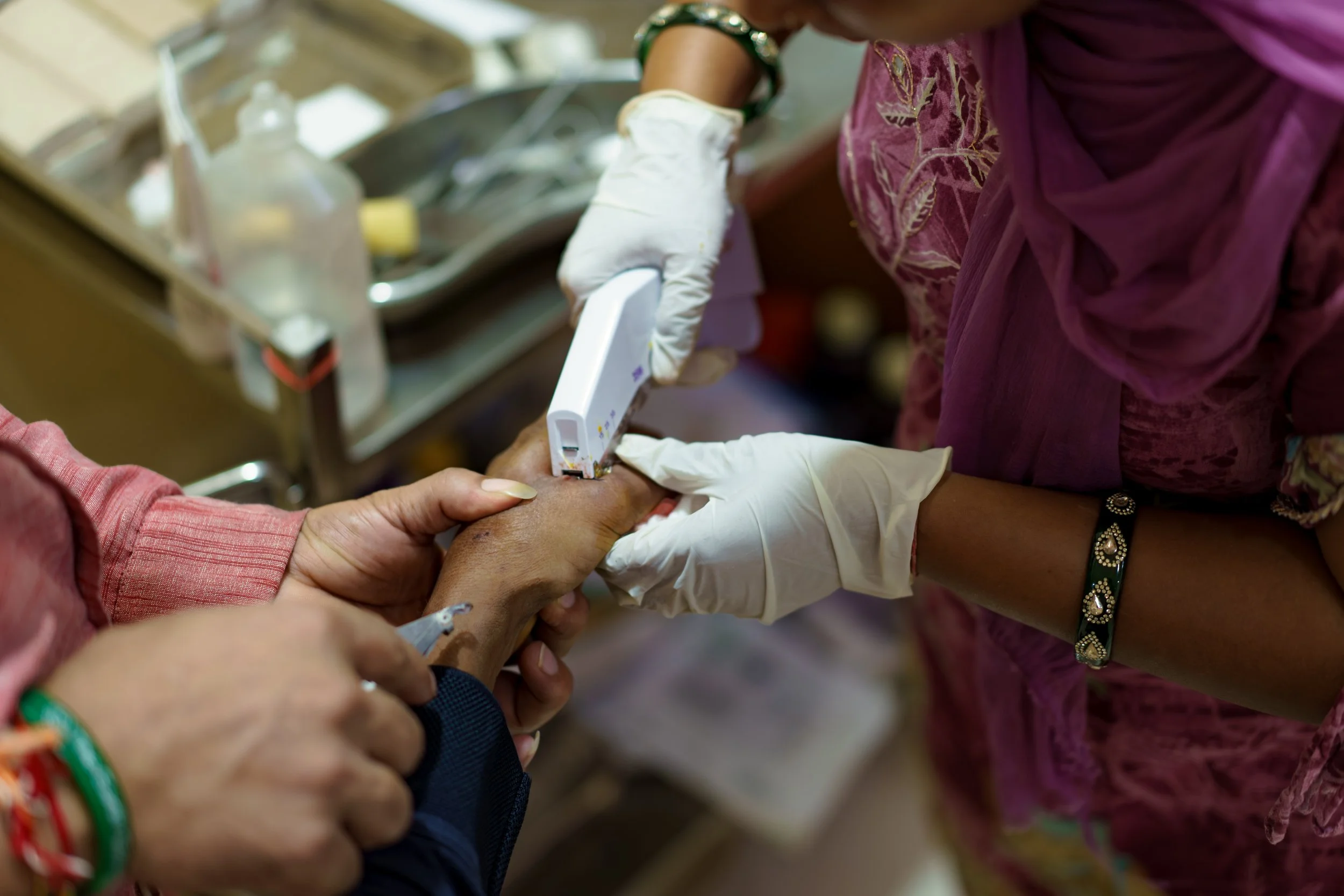 A healthcare worker wearing gloves and a purple saree is drawing blood from a patient's hand using a blood collection device. The patient's hand is supported by another person's hand, and medical supplies are visible in the background.