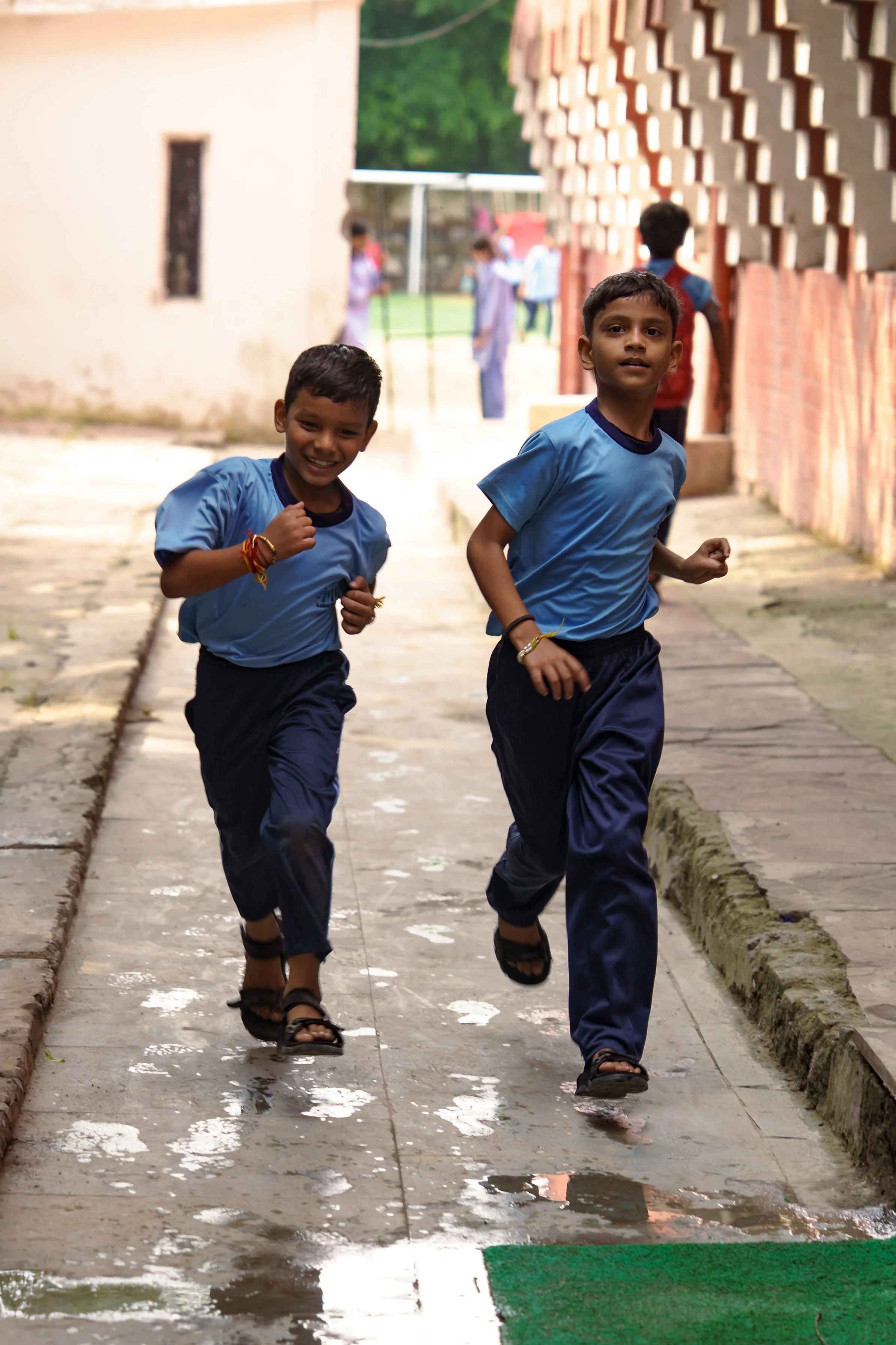 Two boys running on a wet sidewalk, smiling and wearing school uniforms. Other children are visible in the background.