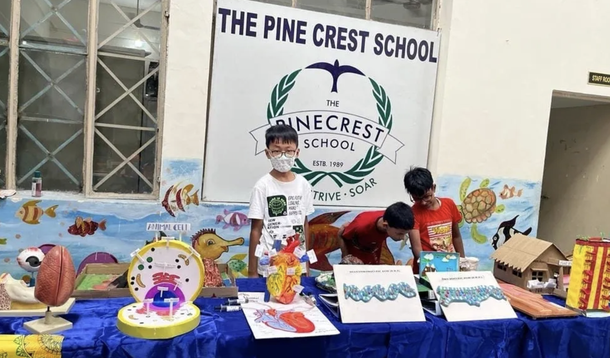 Children at a school fair booth with educational and creative projects, in front of a sign for The Pine Crest School.