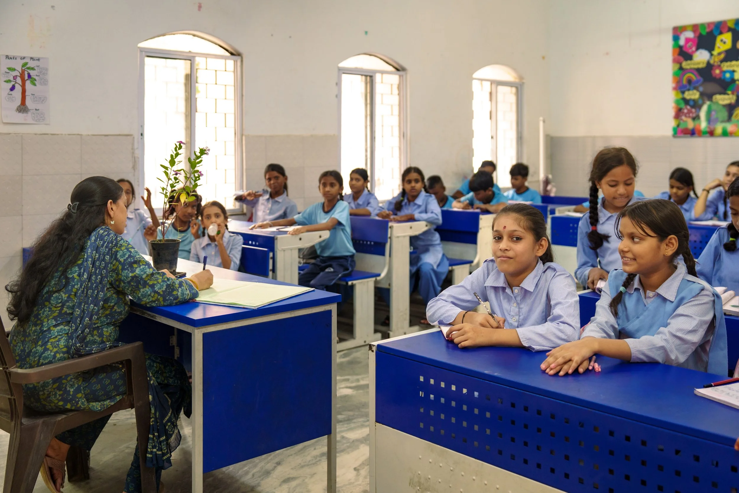 A classroom filled with young students in blue uniforms sitting at blue desks, listening to a teacher who is writing in a notebook, with windows in the background and colorful posters on the wall.