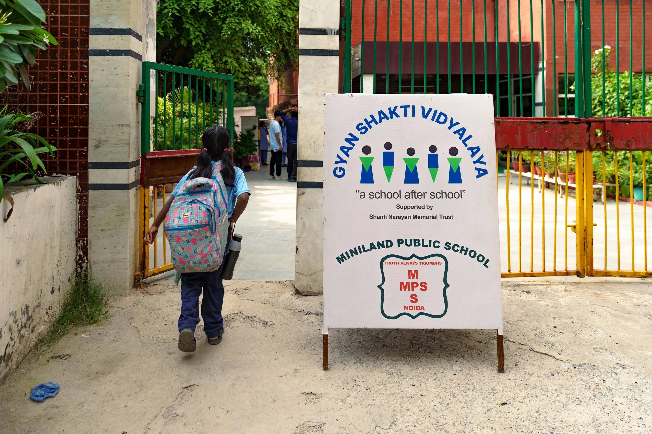 A young girl with a backpack walking towards school through an open gate at Gyan Shakti Vidyalaya, supported by Shanti Narayan Memorial Trust, with a sign providing information about the school.