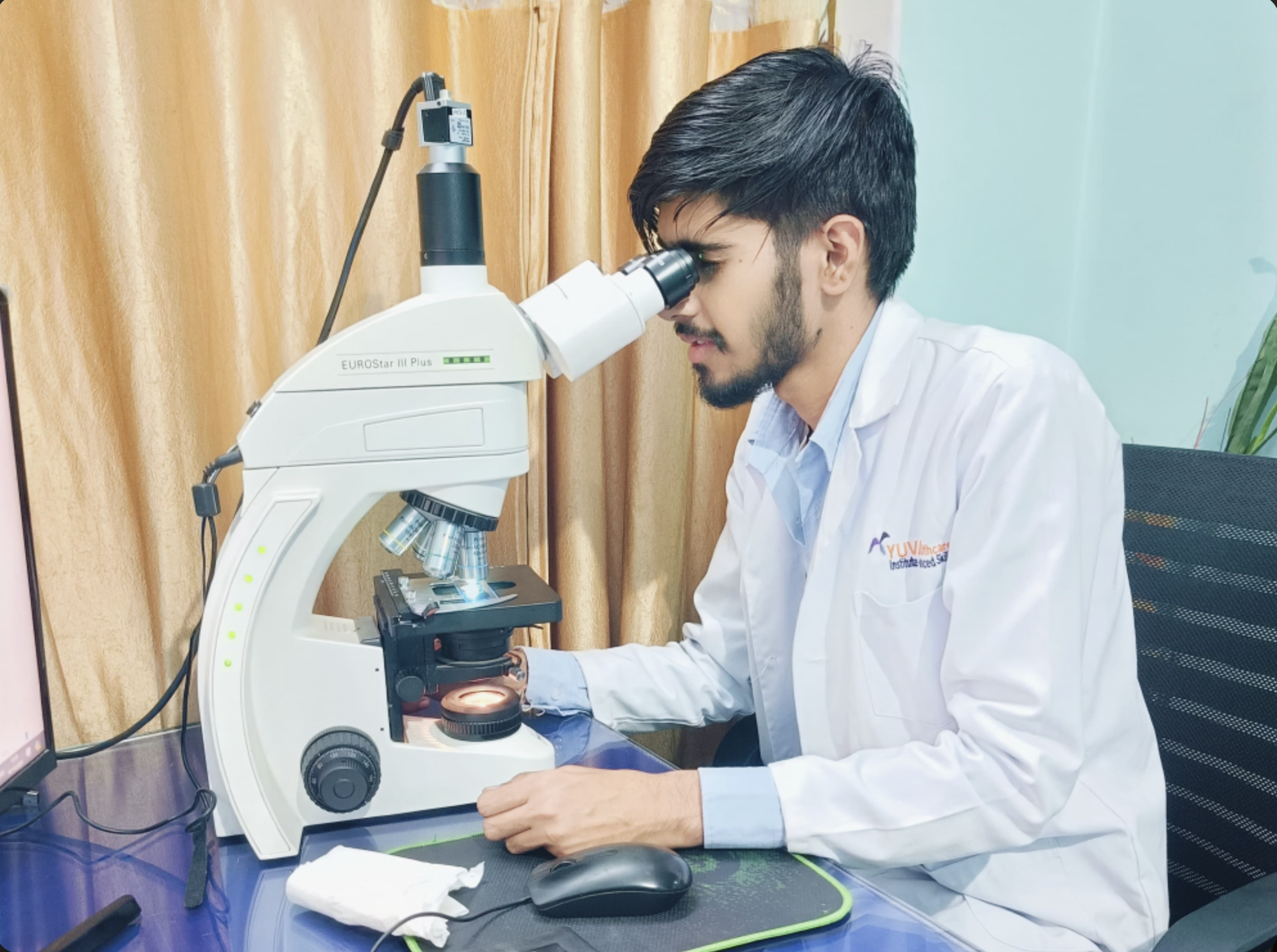 A scientist or researcher looks through a microscope in a laboratory setting. He is seated at a desk with a computer monitor, mouse, and other lab equipment nearby.