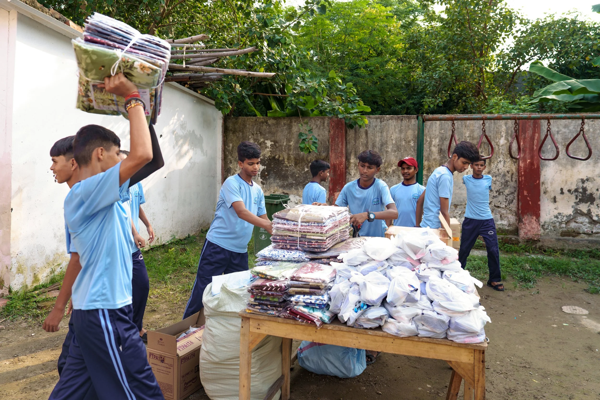 A group of boys in blue shirts collecting and organizing folded clothes and supplies outdoors, with a table holding stacked fabrics and white bags, fencing, and green trees in the background.