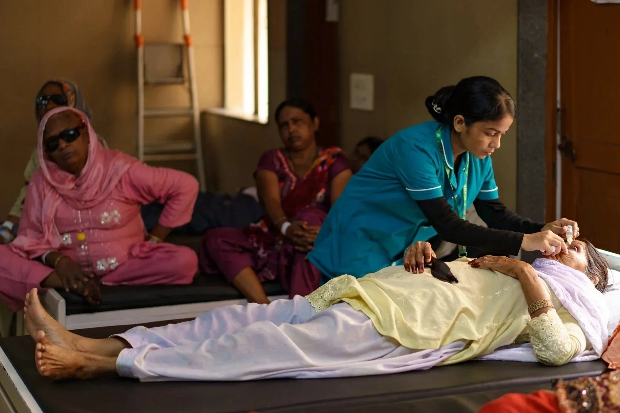 A medical professional performs a procedure on a patient lying on a hospital bed, with three other women sitting nearby and observing in a room with beige walls and a window.