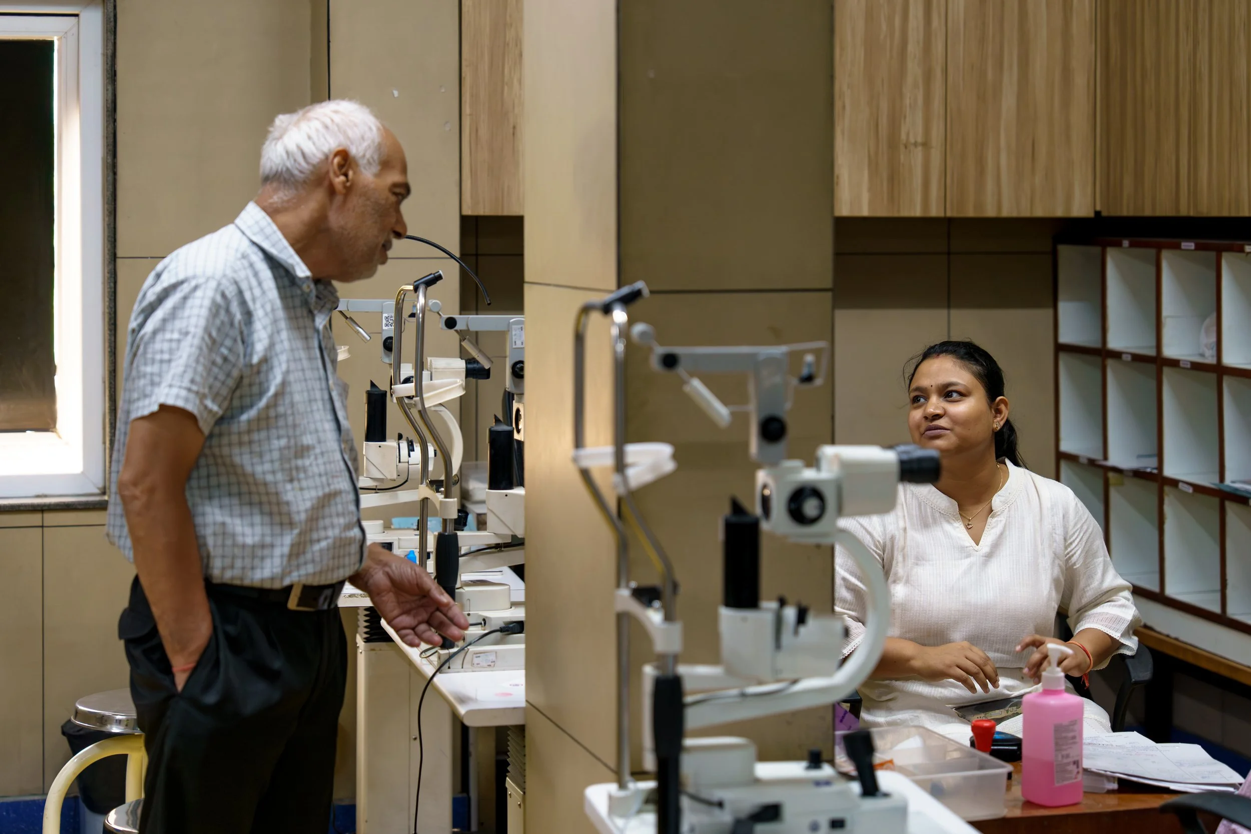 An older man with gray hair and a young woman with dark hair are in a laboratory, engaging in conversation. The man is standing and leaning slightly forward, while the woman is sitting at a table with scientific equipment, including microscopes and b