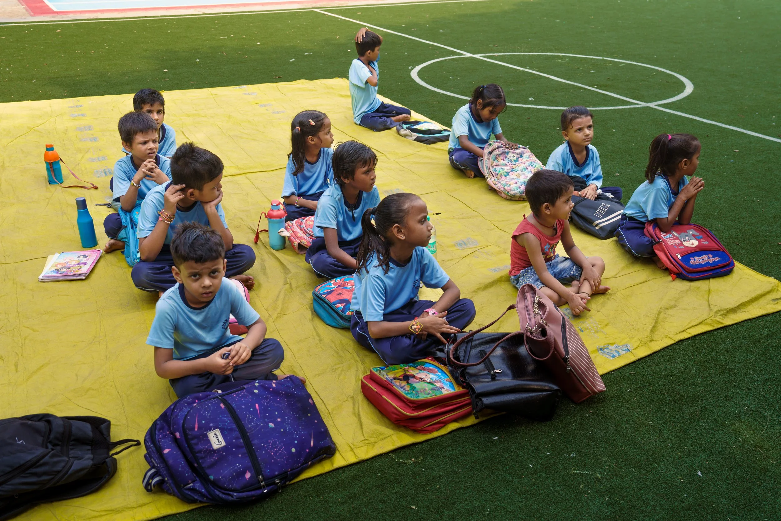 Group of children sitting on a yellow mat on an outdoor sports field with backpacks and water bottles.