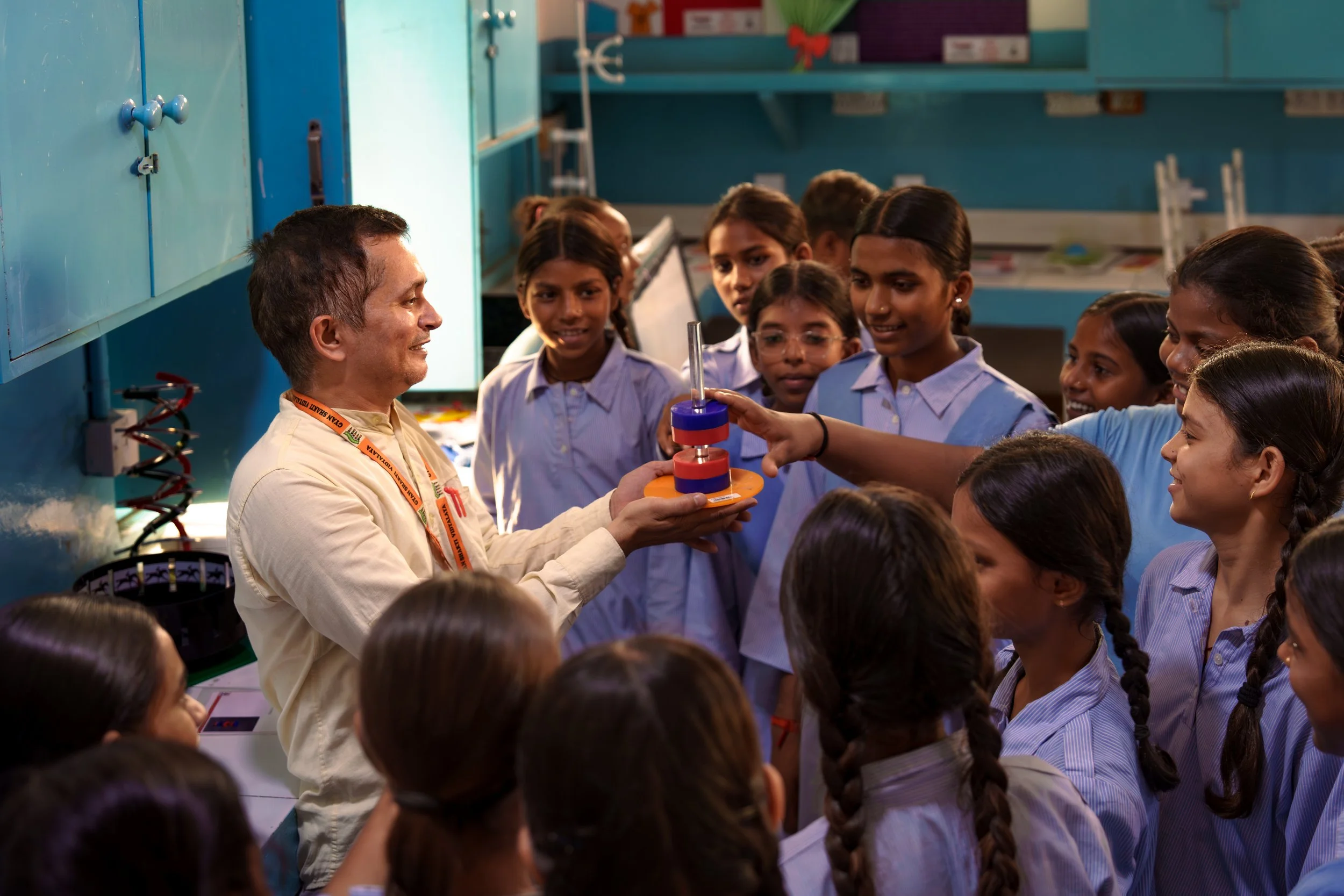 A group of schoolgirls gathered around a teacher in a science classroom, holding a science experiment on a rotating platform with a test tube, with a blue wall and shelves in the background.