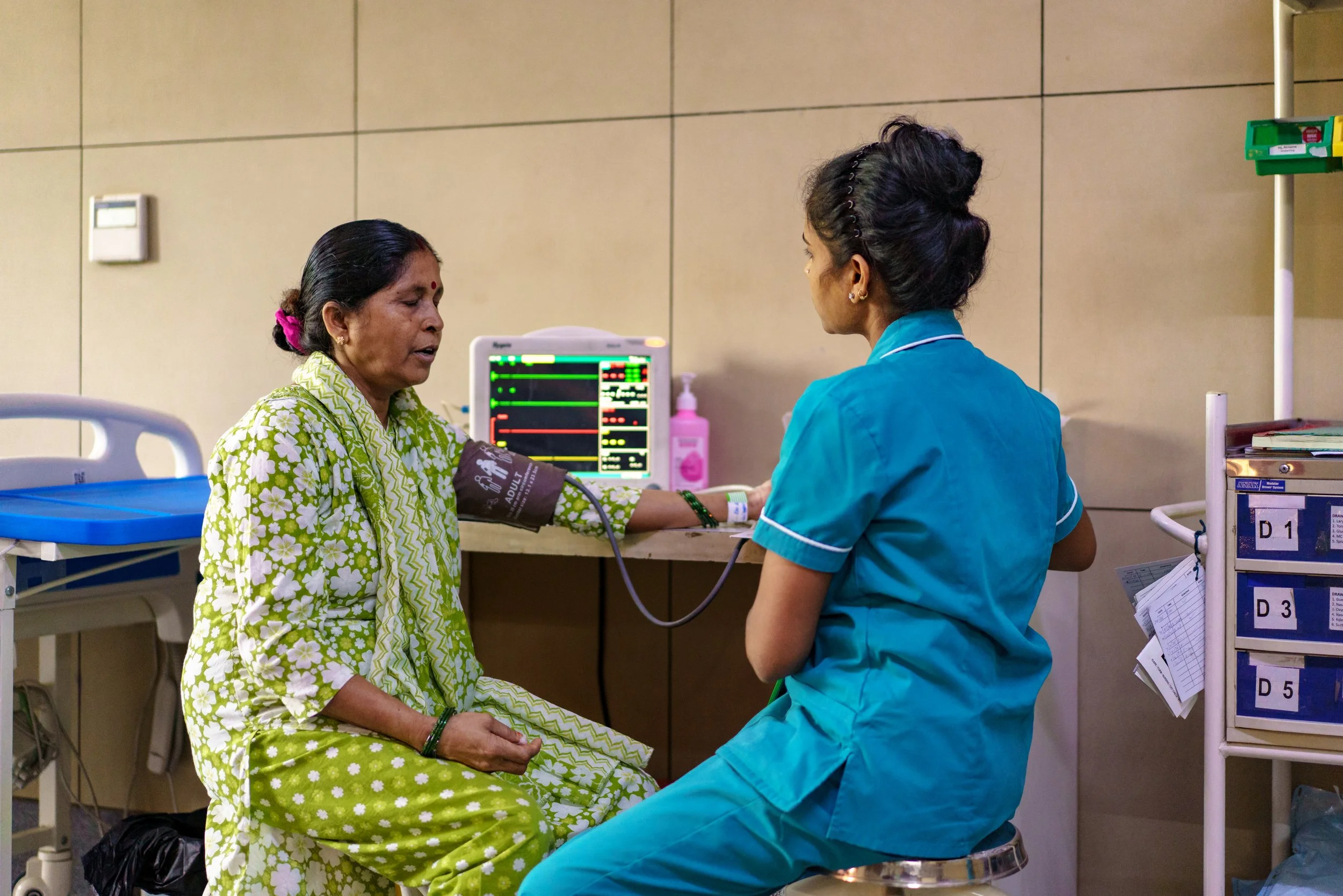 A woman in a green floral hospital gown is having her blood pressure checked by a nurse in blue scrubs in a hospital room.