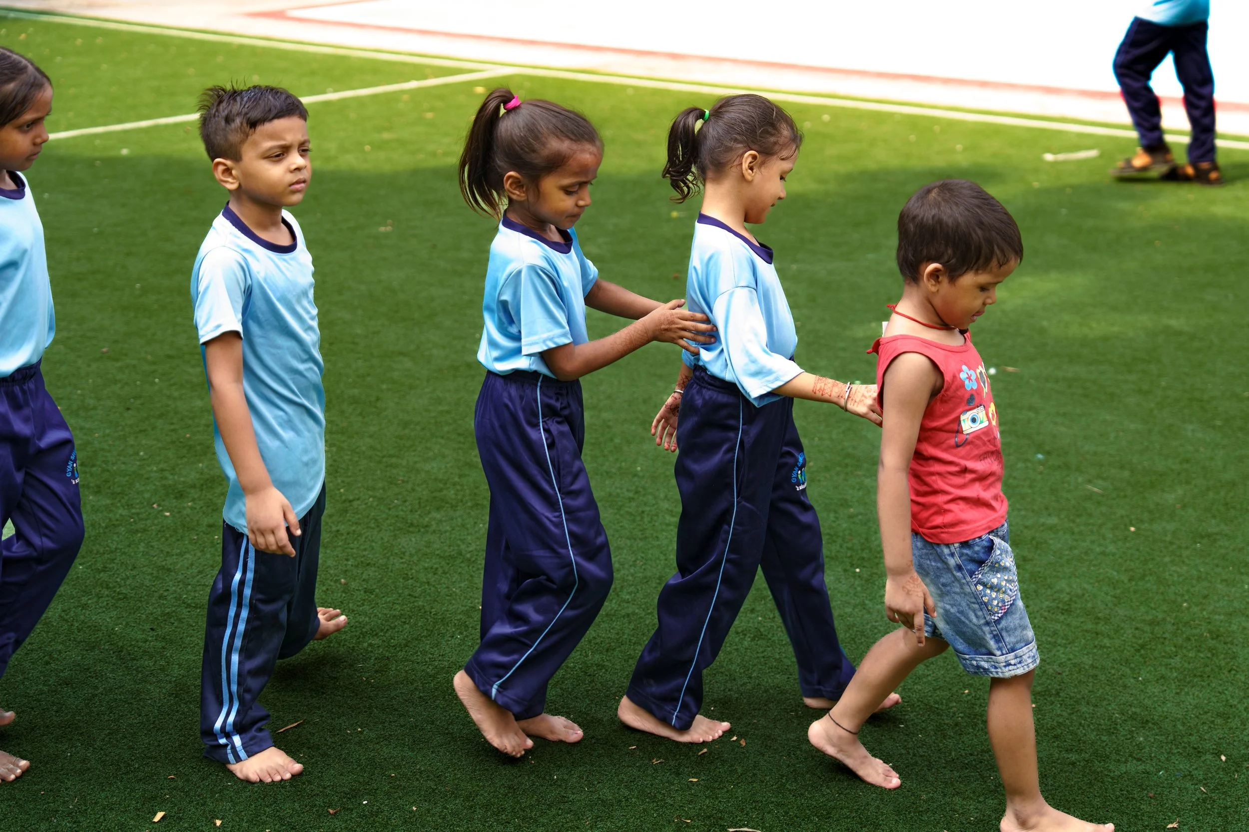 Children walking in a line on a grassy field, holding each other's shoulders, with some children barefoot and dressed in sports uniforms and casual clothes.