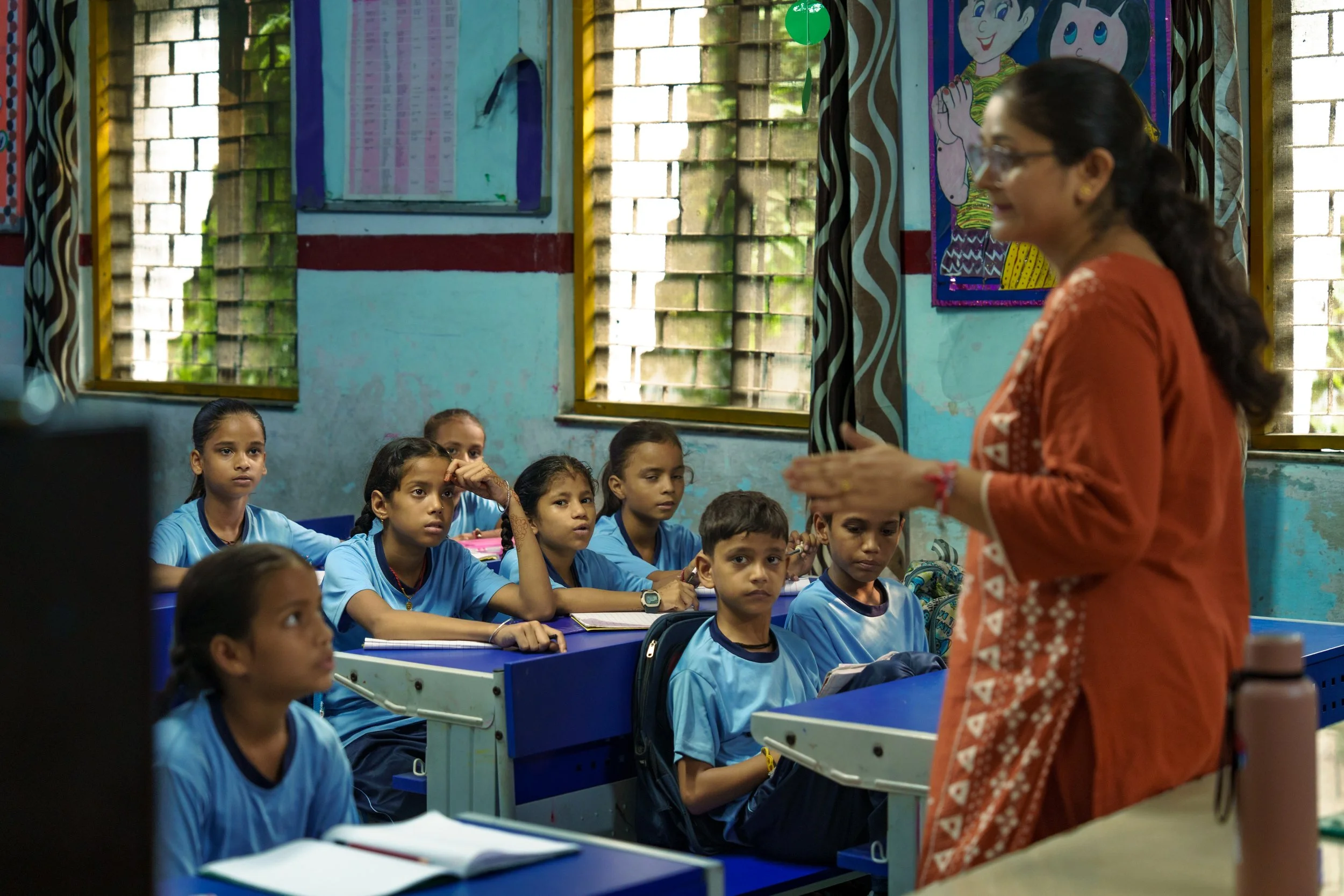 A classroom with young students wearing blue uniforms sitting at blue desks, listening to a teacher. The classroom has large windows with yellow frames, and colorful posters on the walls, including cartoon characters. The teacher, an adult woman wear
