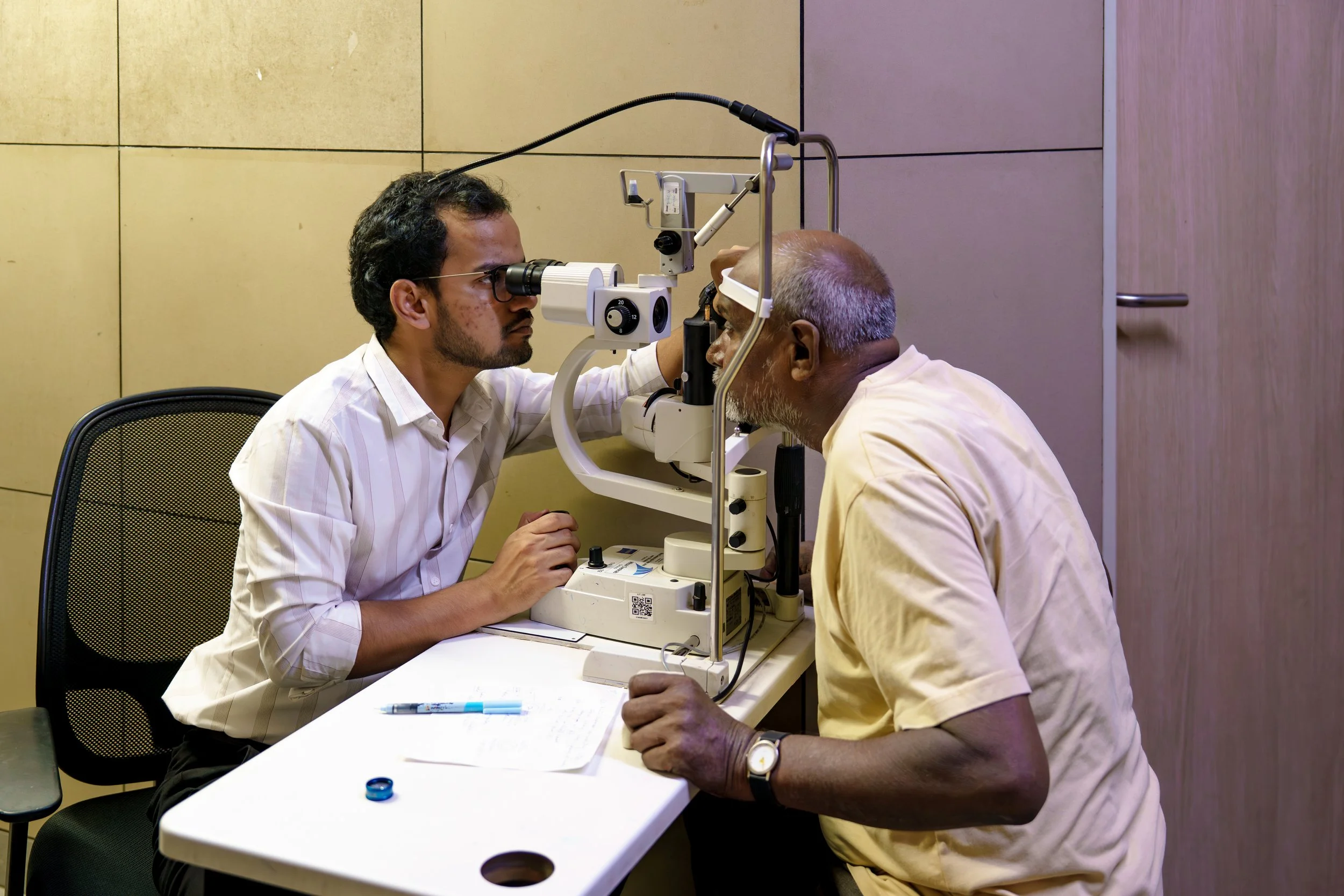 An eye doctor examines an elderly man using a slit lamp in a clinical setting.