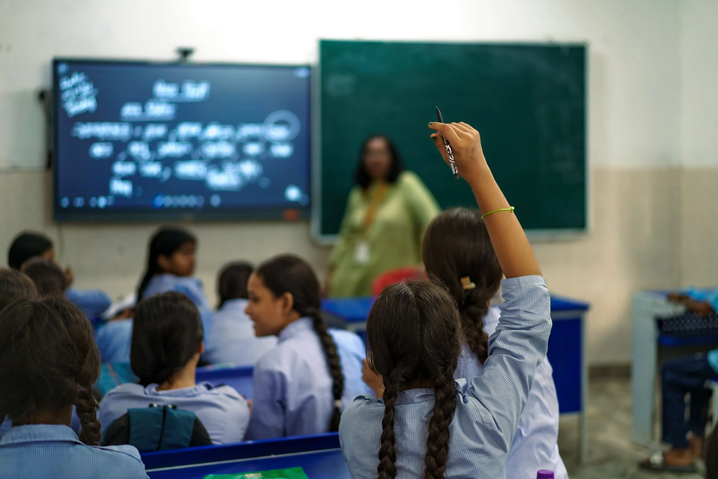 A classroom scene with students seated and a hand raised of a student in the foreground, and a teacher standing in front of a chalkboard and a digital screen displaying writing in the background.