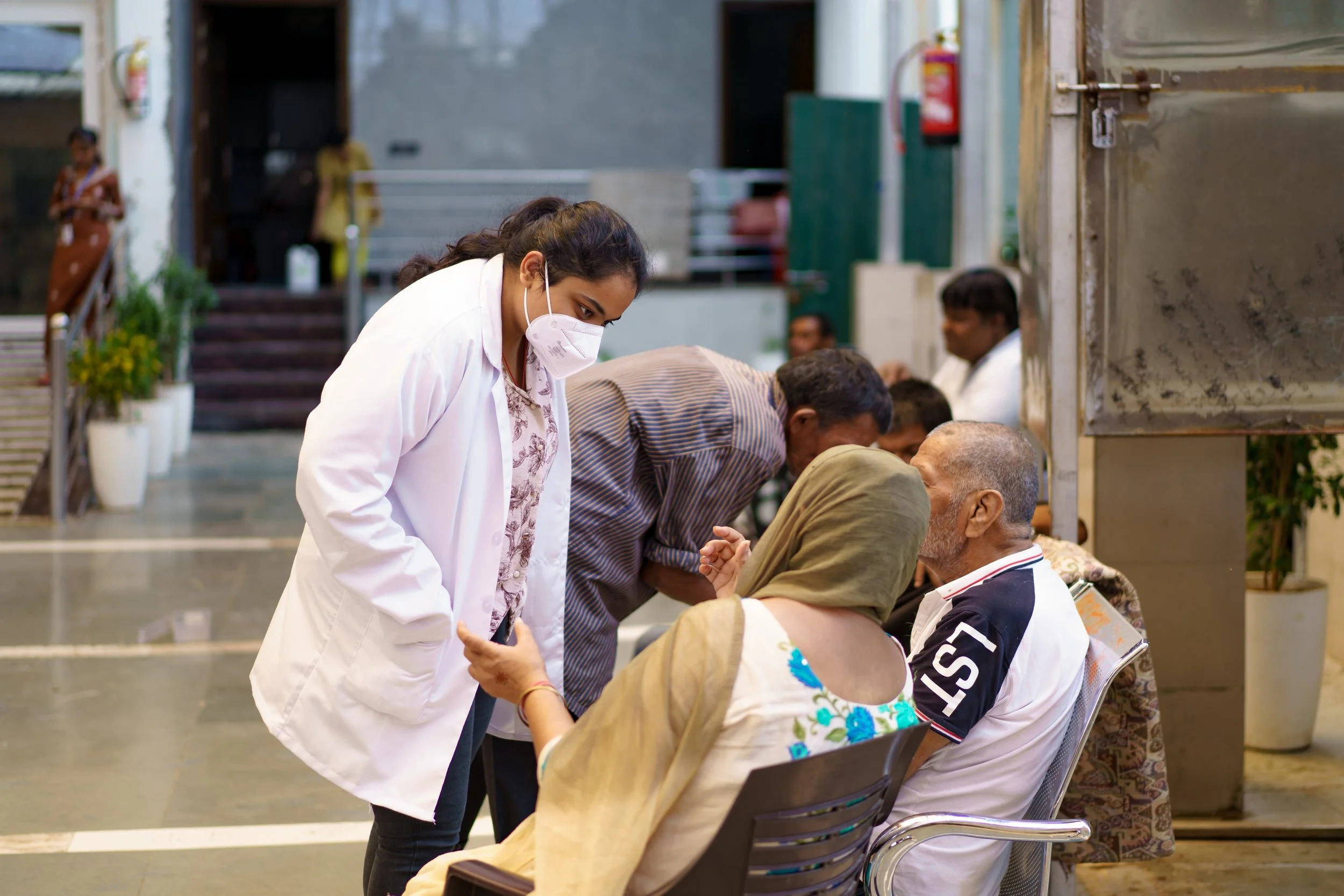A woman wearing a mask and white coat is standing in a line, greeting and shaking hands with seated elderly individuals. Other people are in the background in an indoor setting.