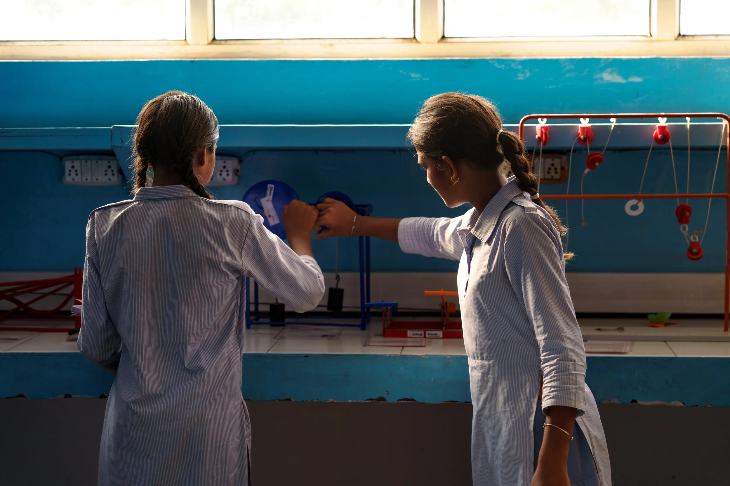 Two girls with braided hair wearing school uniforms in a classroom, with a blue wall and large windows in the background.