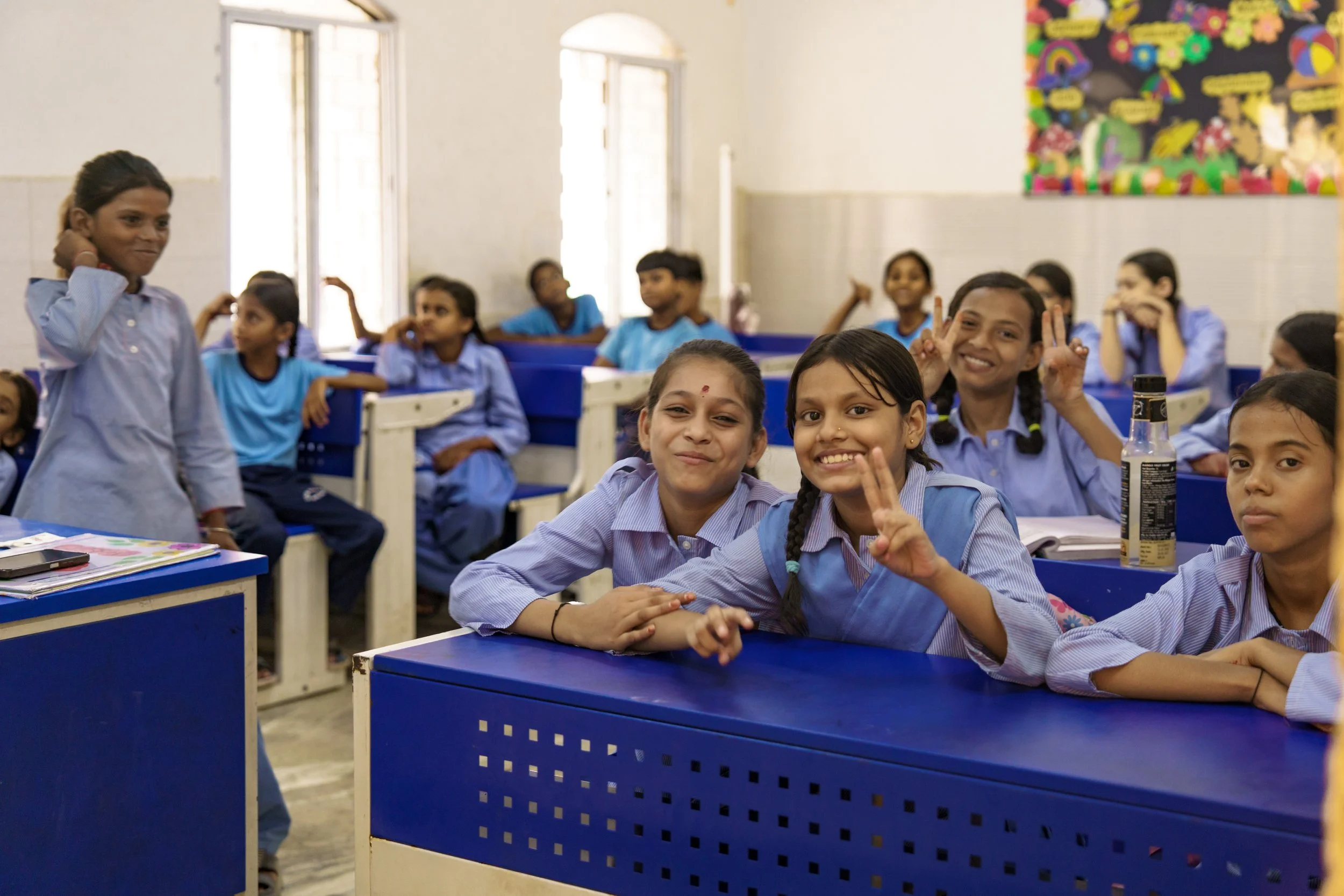 Classroom full of smiling students in blue uniforms, some making peace signs, sitting at blue desks with a colorful bulletin board on the wall.