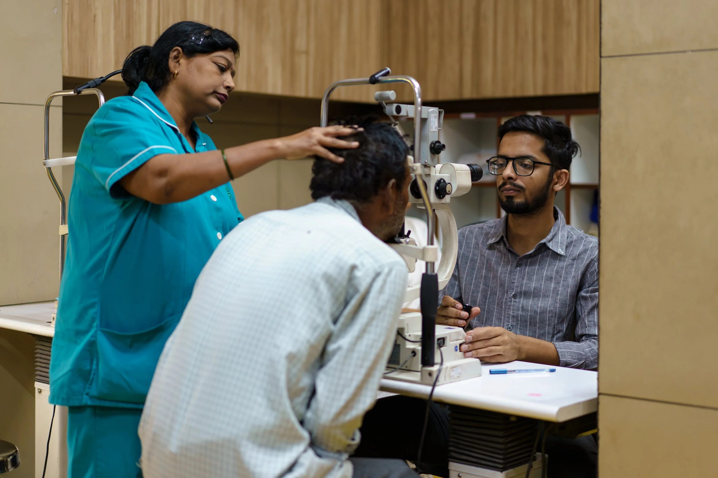 An eye doctor examines a patient using specialized ophthalmic equipment while a woman assistant looks on, in a clinical setting.