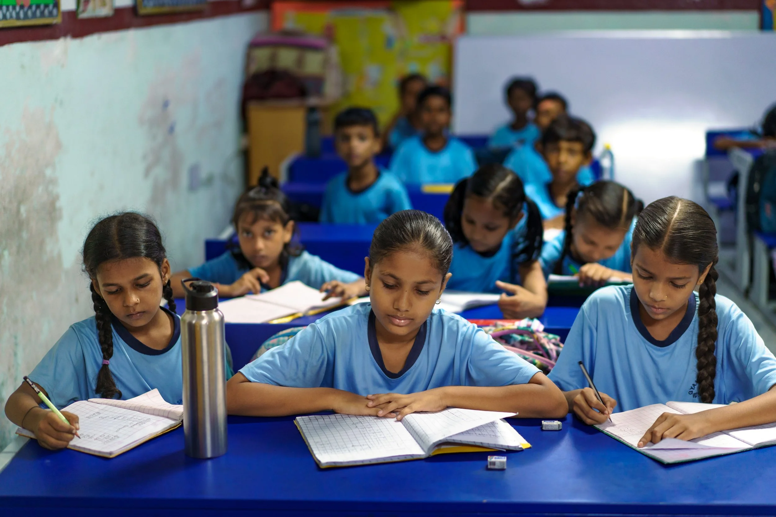 Elementary school classroom with young students focused on their notebooks at desks, wearing light blue uniforms.
