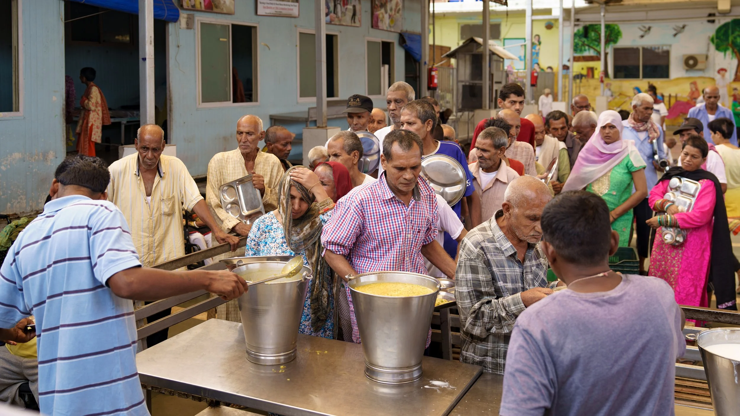 People lined up at a food distribution event, with large pots of food and serving trays, indoors with colorful walls and murals.