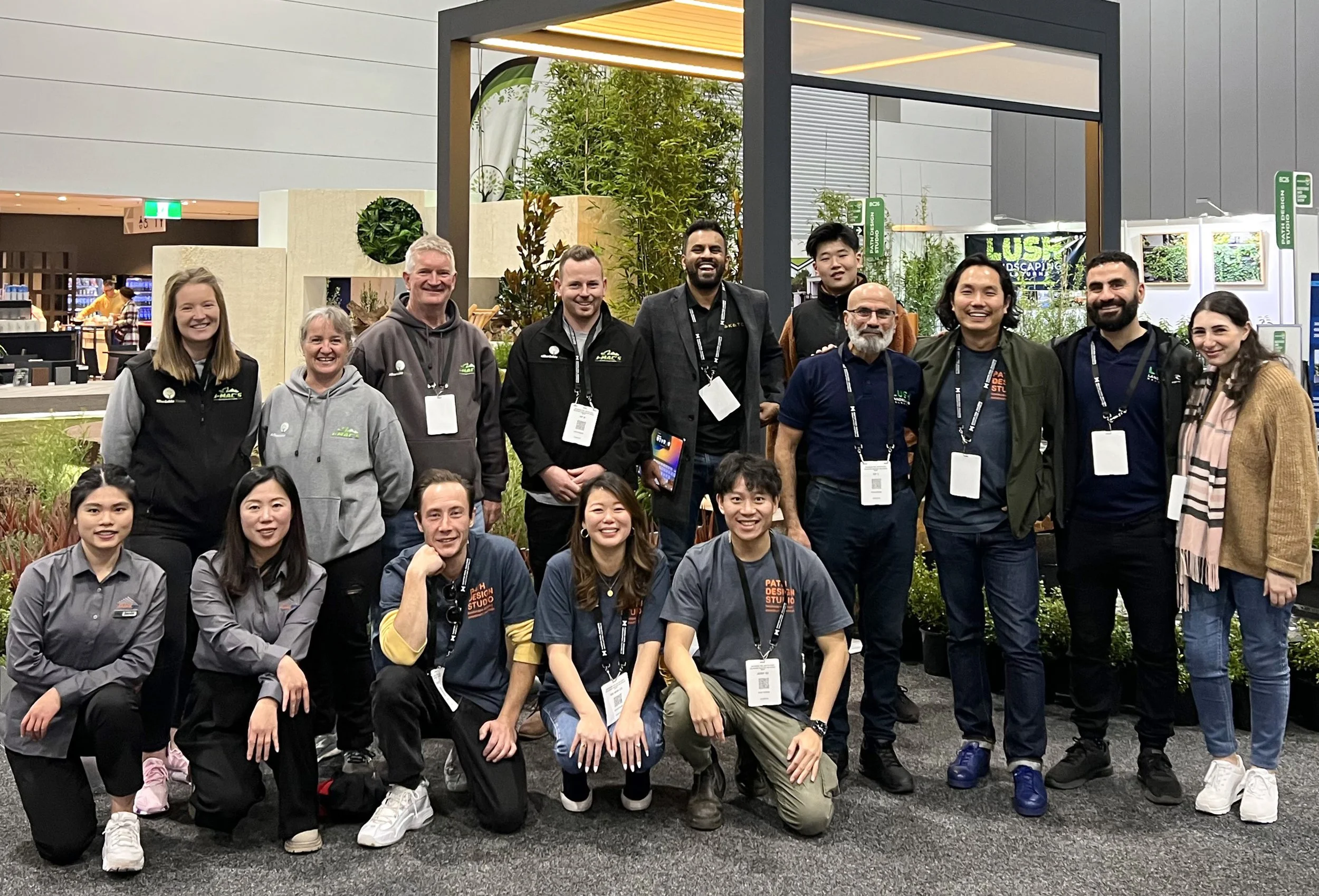 A group of diverse people standing together at an indoor event, smiling for the camera, with some people seated in front. The background shows a booth with greenery and a sign that says "LUSH."