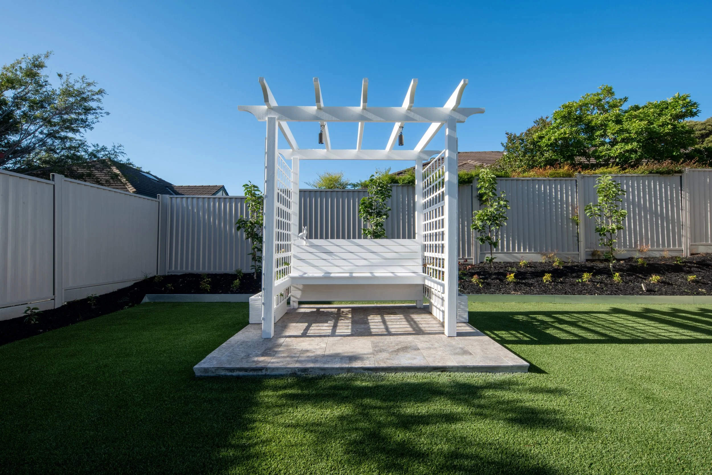 A white outdoor gazebo with lattice sides in a backyard, surrounded by a grass lawn, with a tall fence, trees, and houses in the background under a clear blue sky.