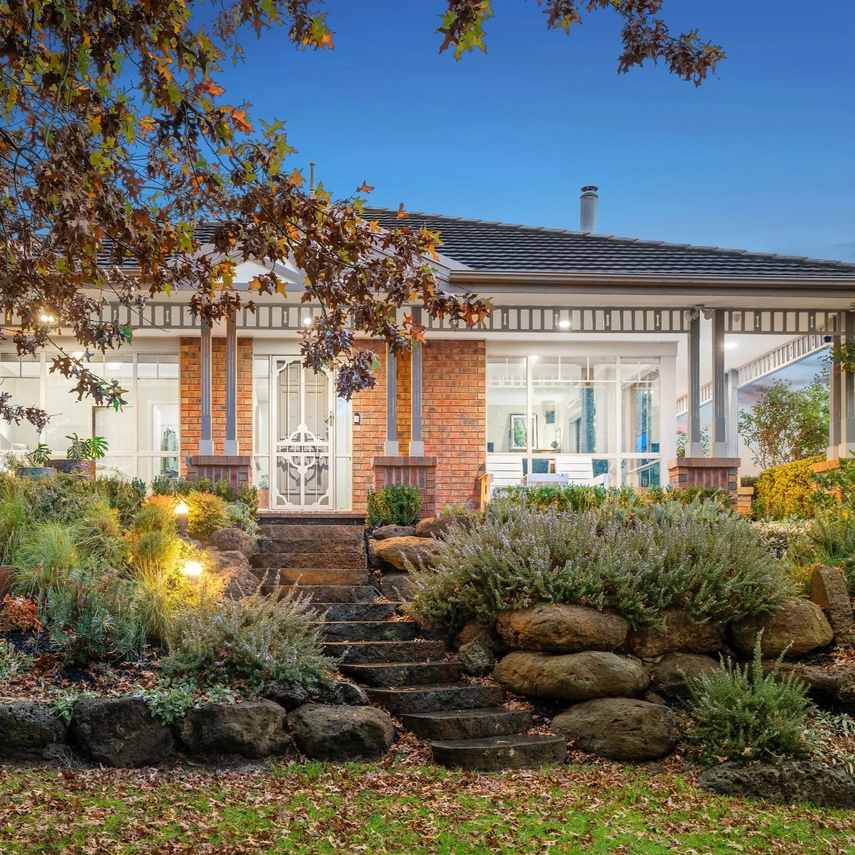 Front view of a house with a garden and stone staircase leading up to the entrance. The house has brick walls, large glass windows, a porch with railing, and a sloped tiled roof. The garden is landscaped with plants, bushes, rocks, and pathway lighting. There are trees with leaves of mixed green and autumn colors, and the sky is clear and blue.