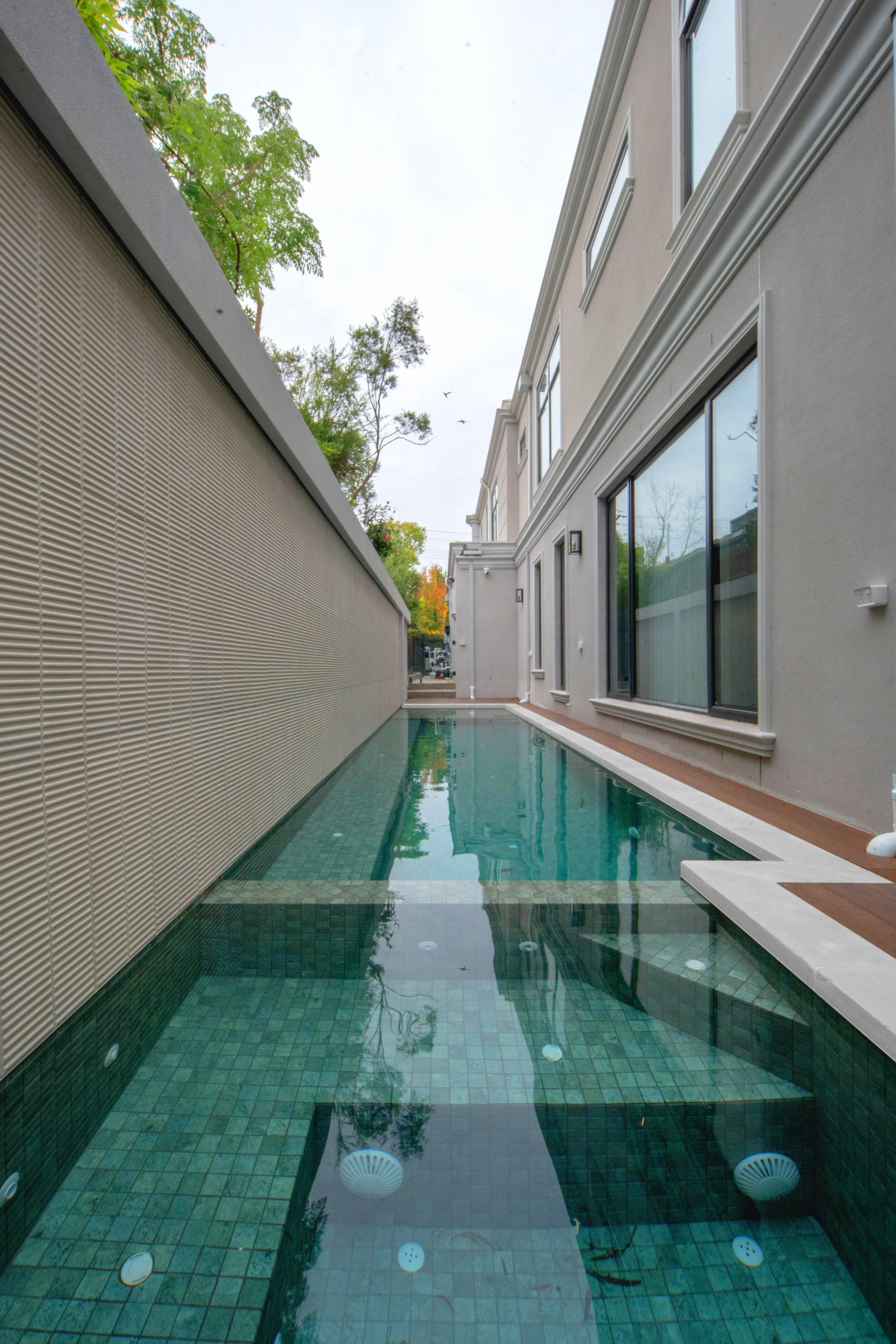 A modern swimming pool situated against a beige residential building with large windows and white trim. The pool has a tiled bottom with jets installed, and is surrounded by a wooden deck. Green trees are visible in the background, and the sky is ove