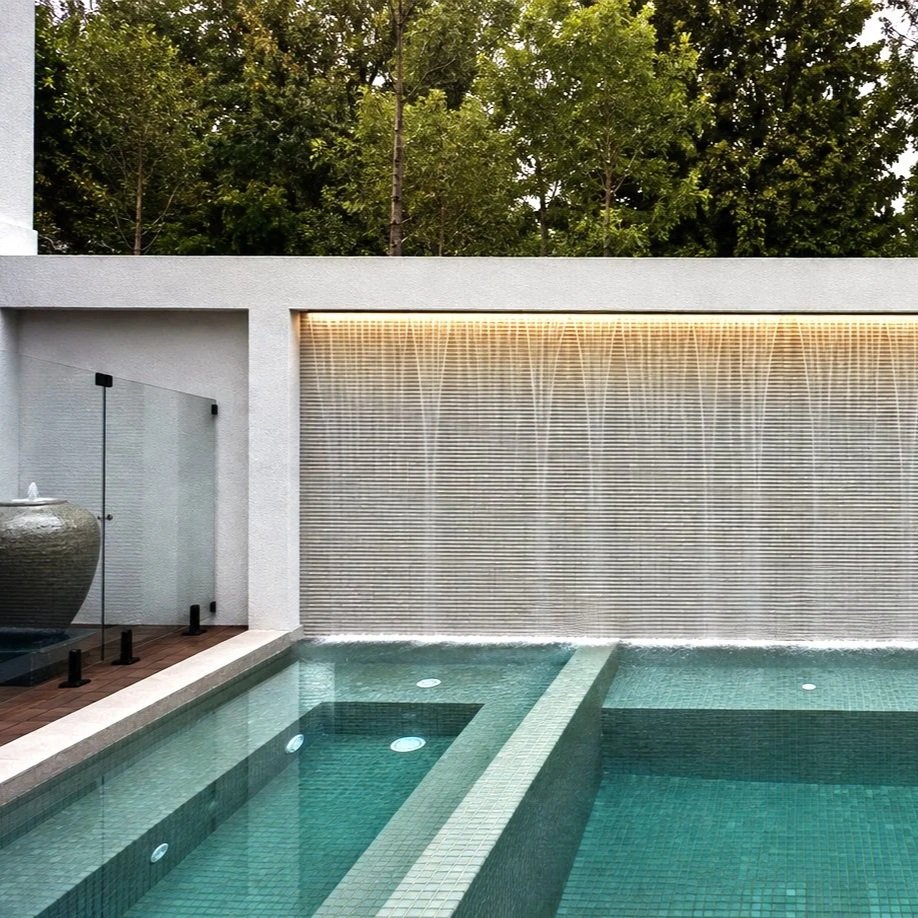 Modern backyard water feature with a water wall, water jets, and a swimming pool, surrounded by green trees.