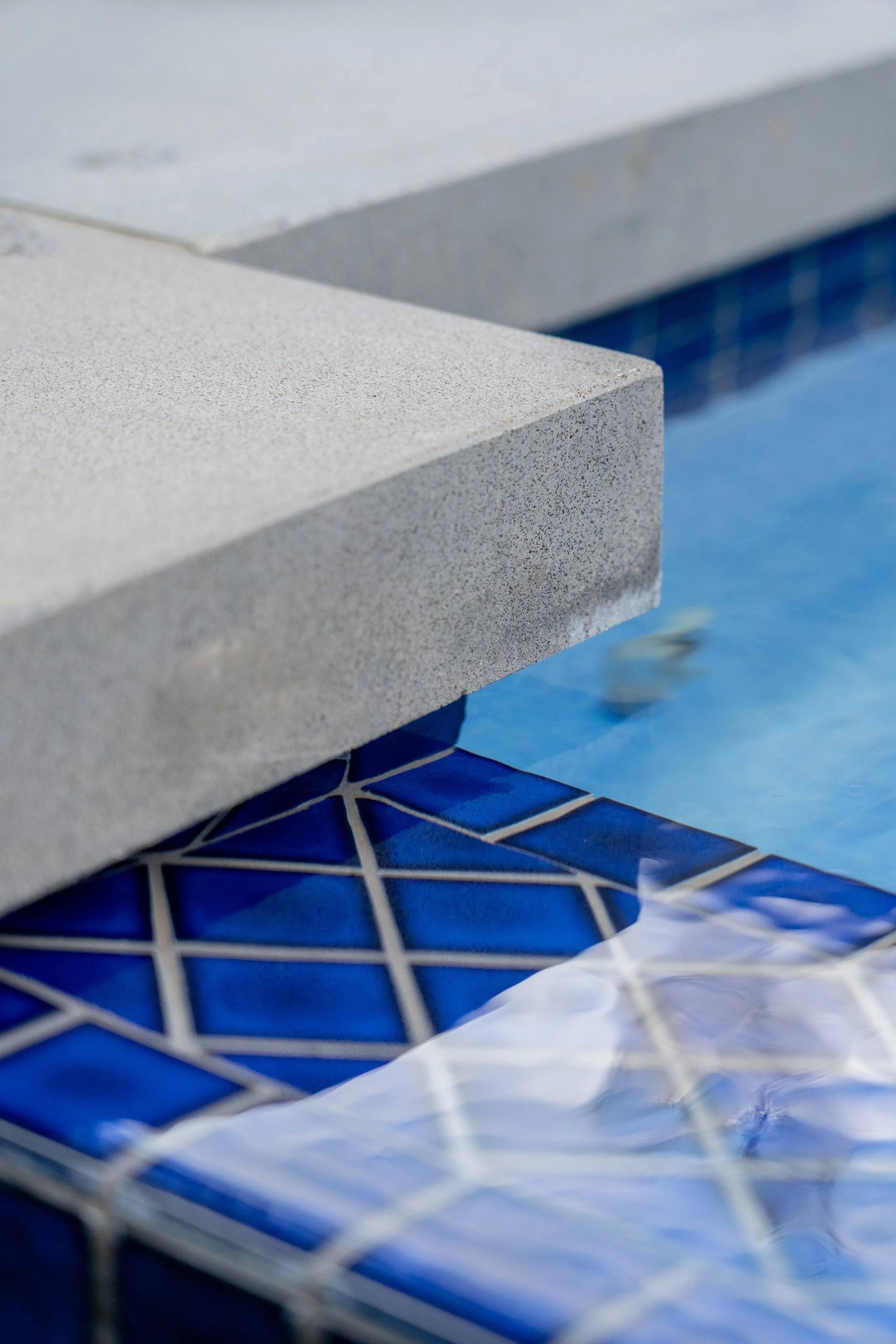 Close-up view of a swimming pool's edge with a blue mosaic tile border and concrete coping, with a portion of the pool water visible.
