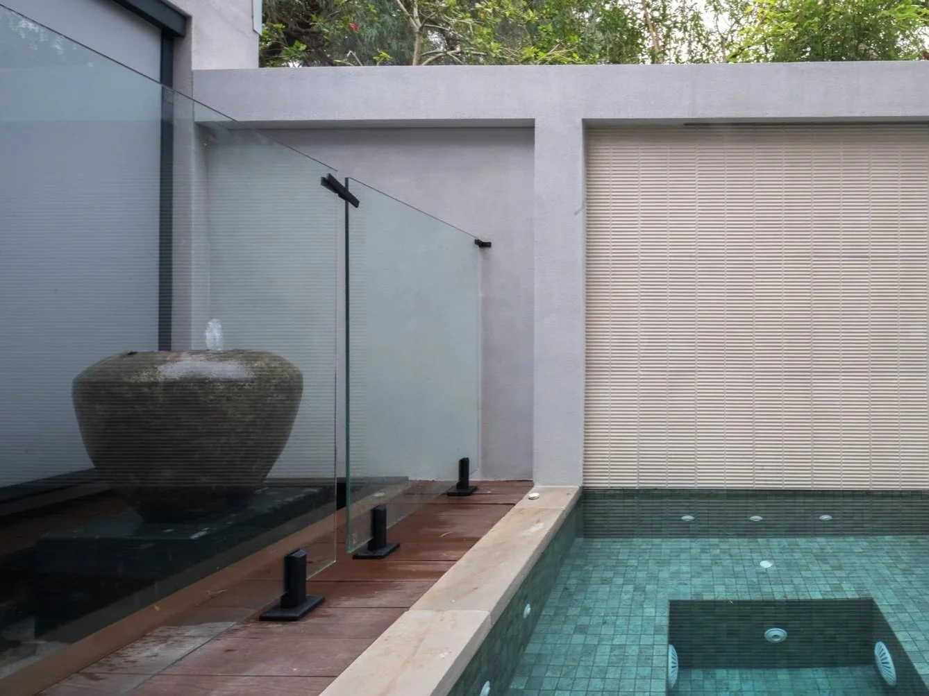 Modern outdoor pool area with a glass snow fence, a large stone fountain, and a beige roller shutter beside the pool, set against a backdrop of trees and a building with concrete and gray walls.