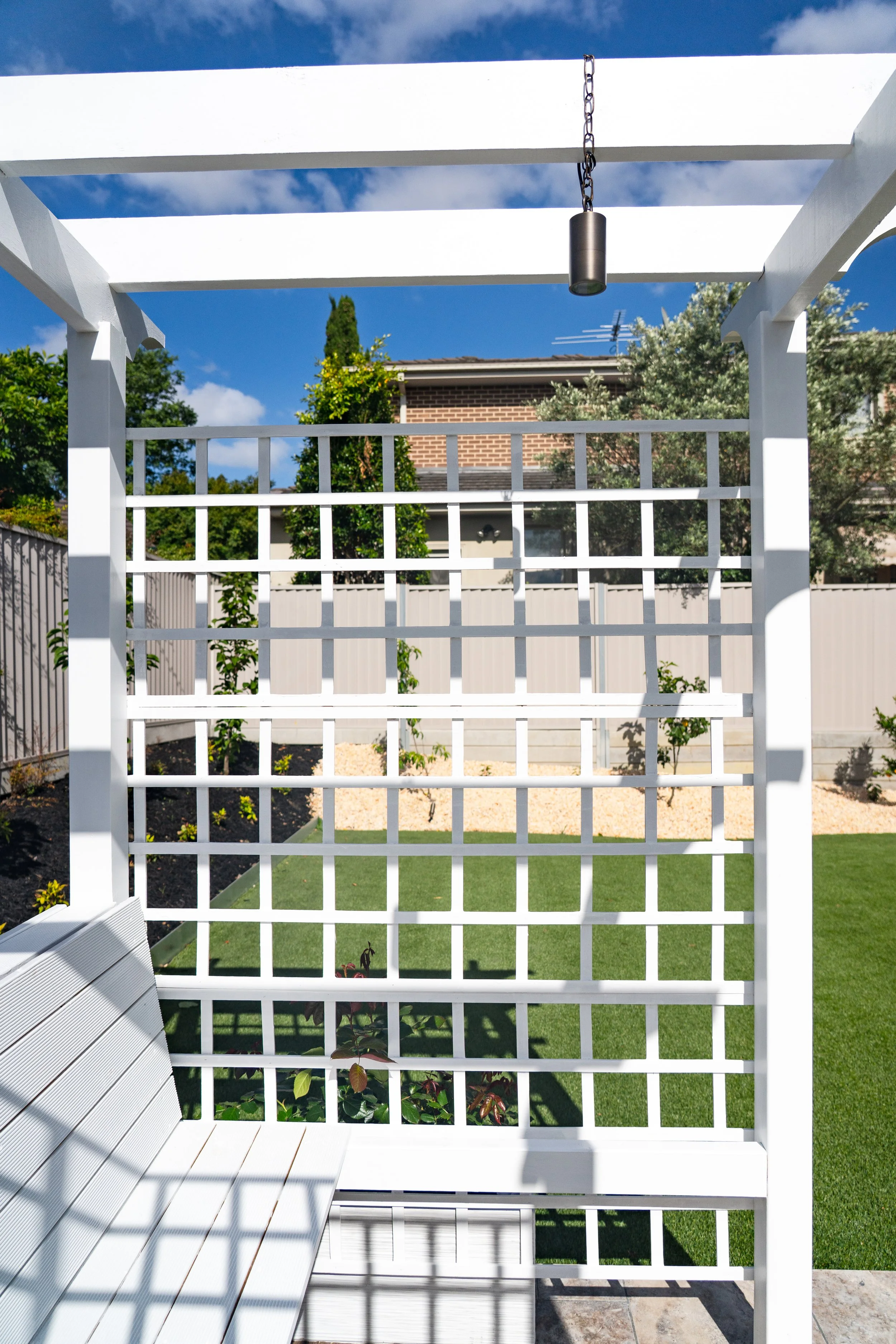 The white timber pergola under the sunlight, creating a partly shaded seating space standing on the lawn, with rose climbing at the rack.