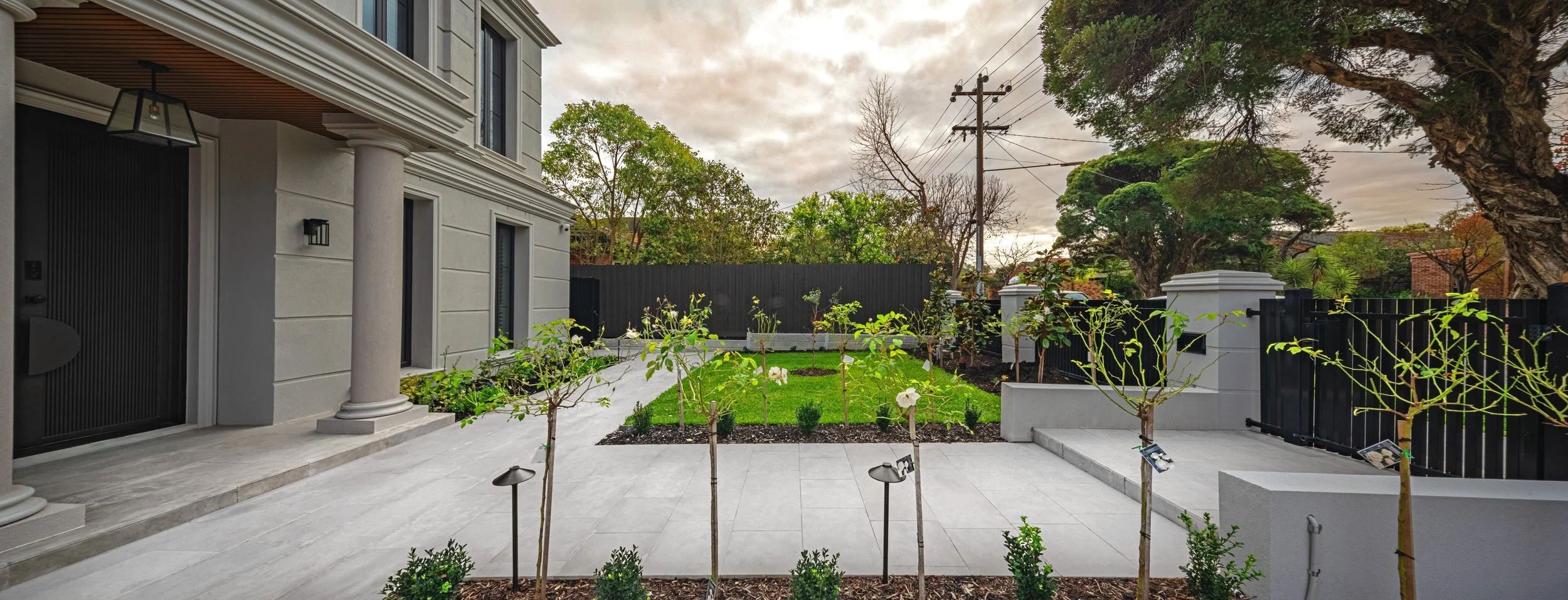 A modern house with a front yard, small trees, and a black fence, with a cloudy sky overhead.