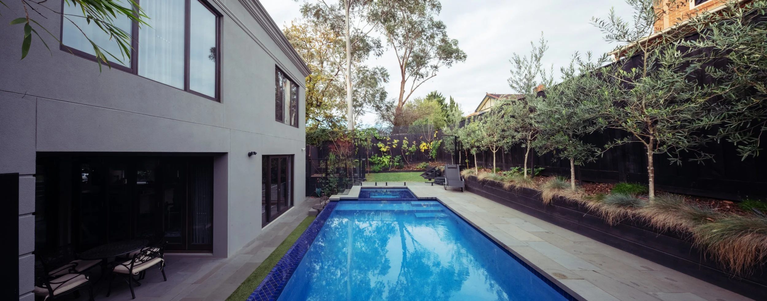 A backyard with a rectangular swimming pool, surrounded by outdoor seating, trees, and a modern house with large windows.