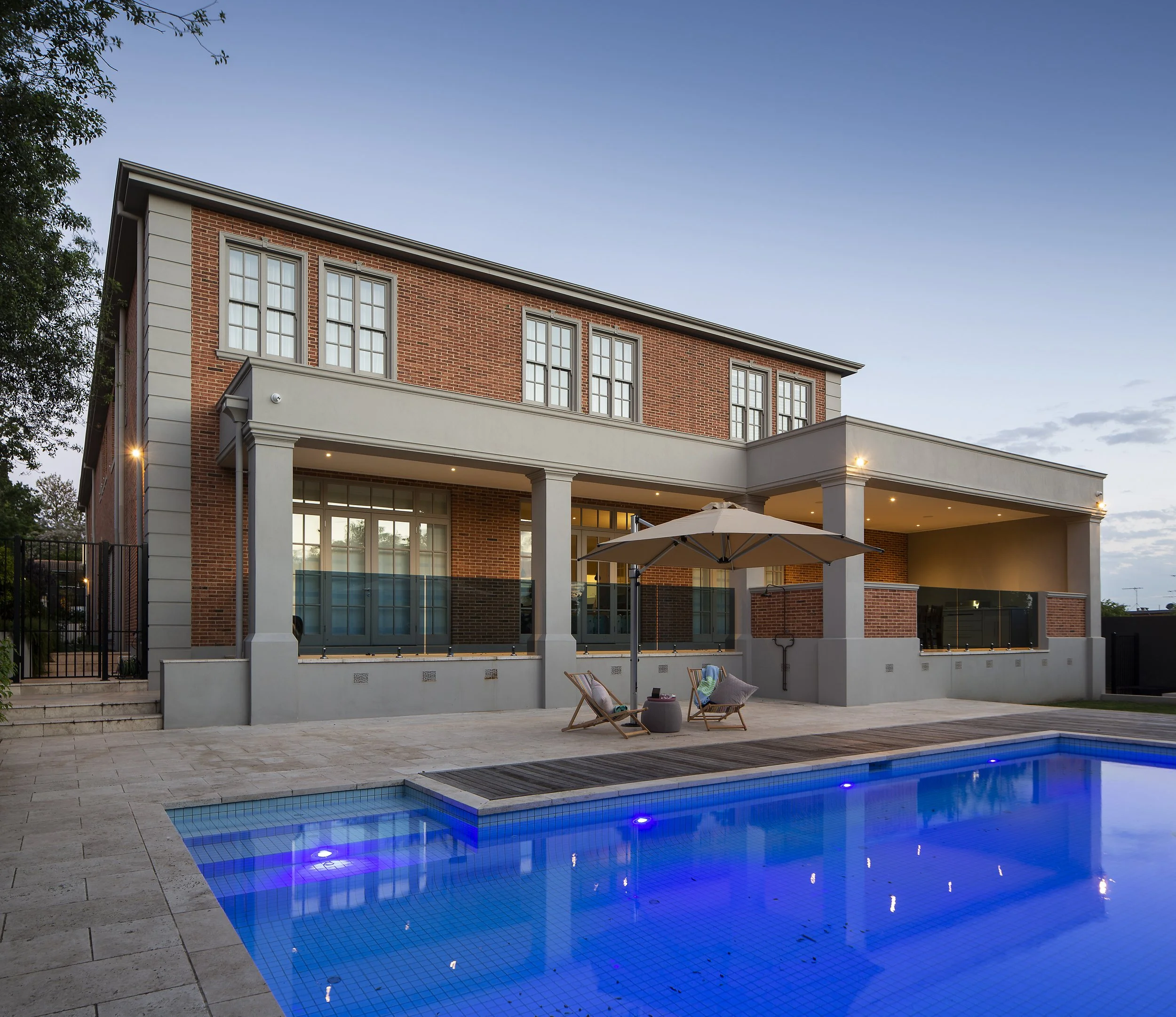 A swim pool on timber decking at the backyard of a mansion, surrounding by hedges of vegetation, showing off a high class life.