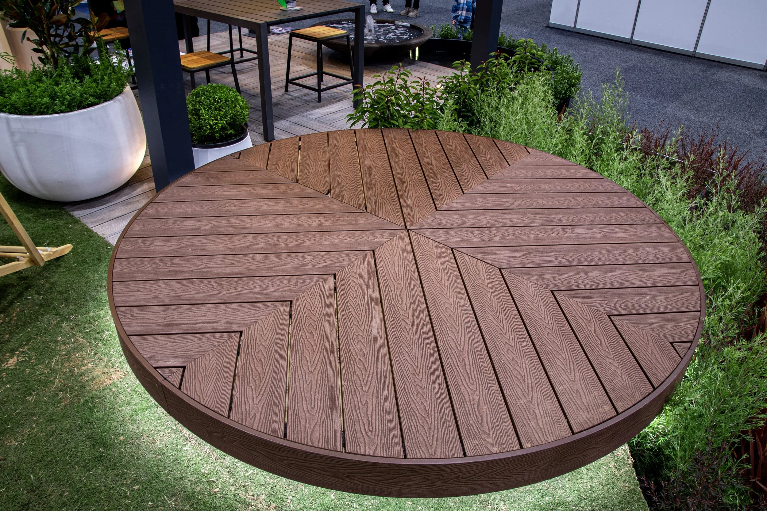 Round wooden table outdoors with greenery around, part of a patio setting with chairs and plants.