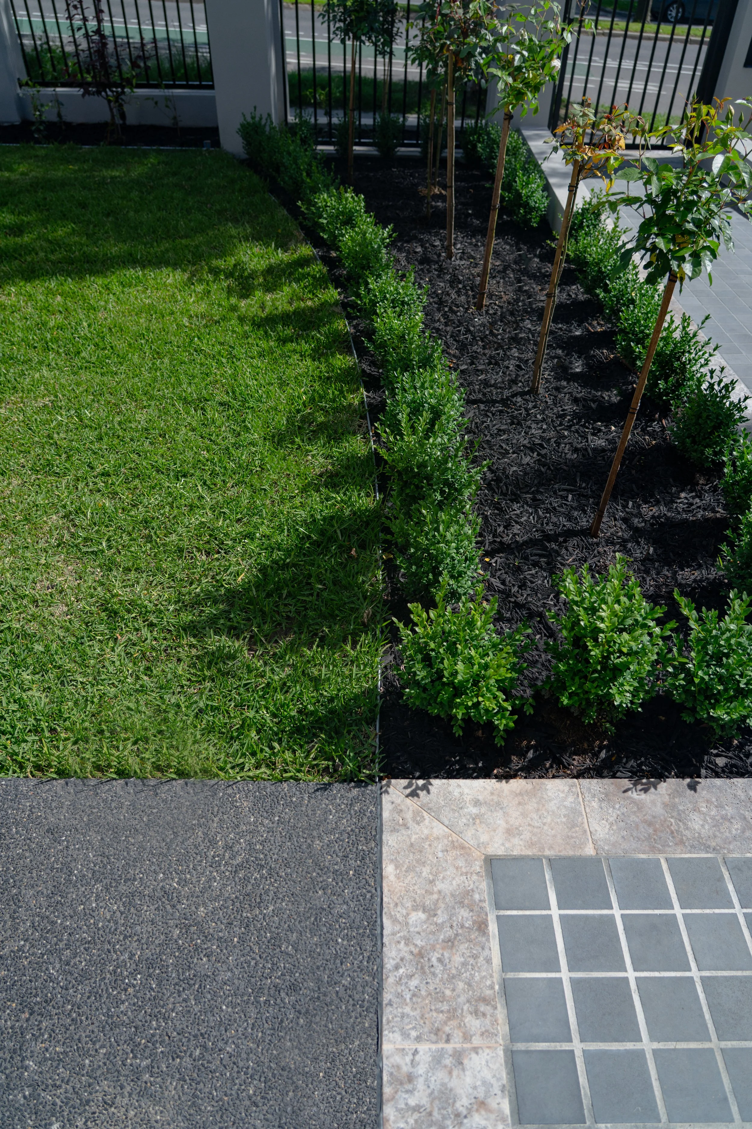 Close-up of a colorful outdoor area with a green grassy lawn, a garden bed with small shrubs and trees, and a tiled pathway, with a black metal fence and street visible in the background.