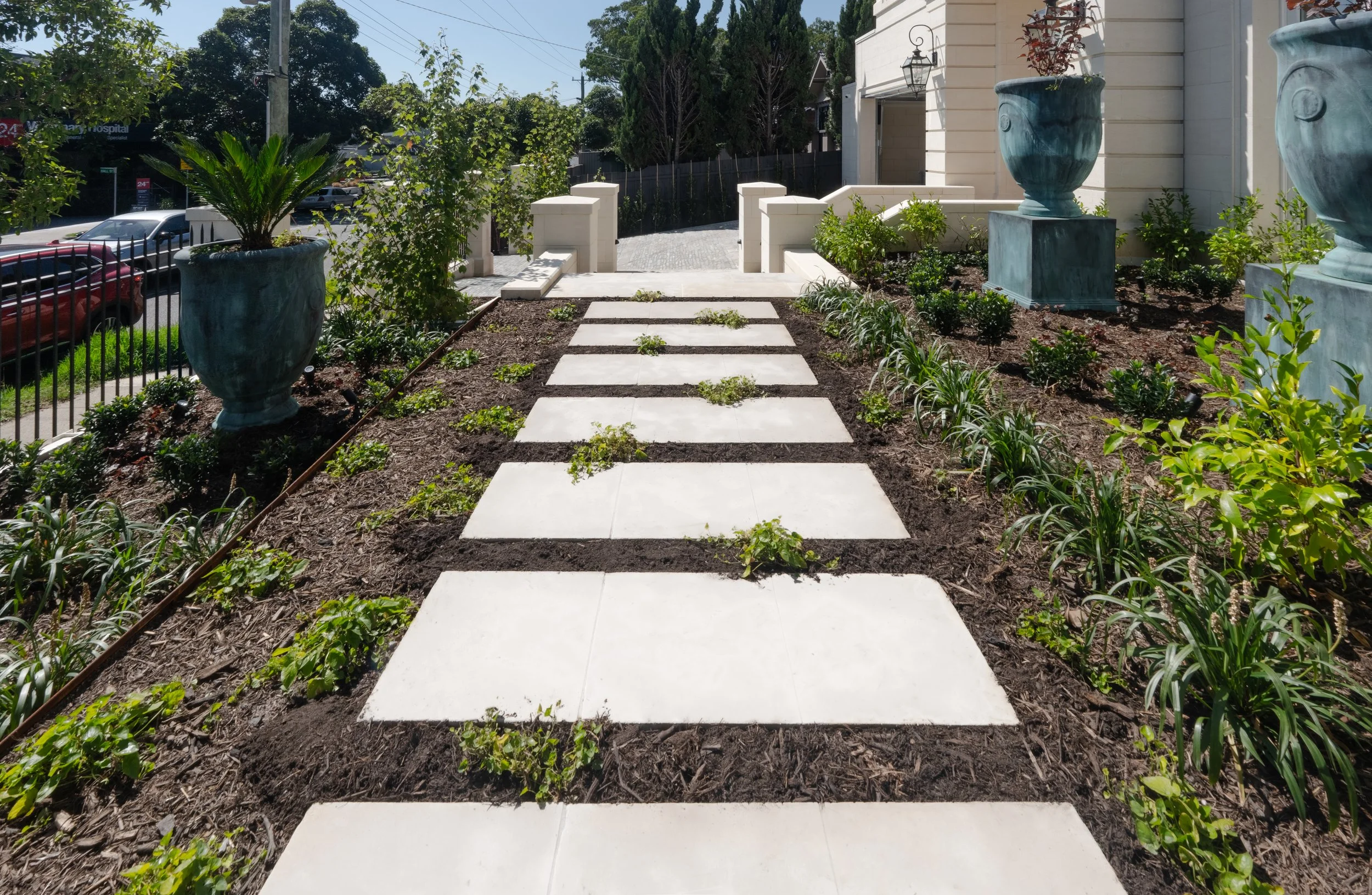 Raised garden bed with different layers of plantations in different height of garden bed, together with the decorative urn and stepping stones creating a high-end looking front garden.