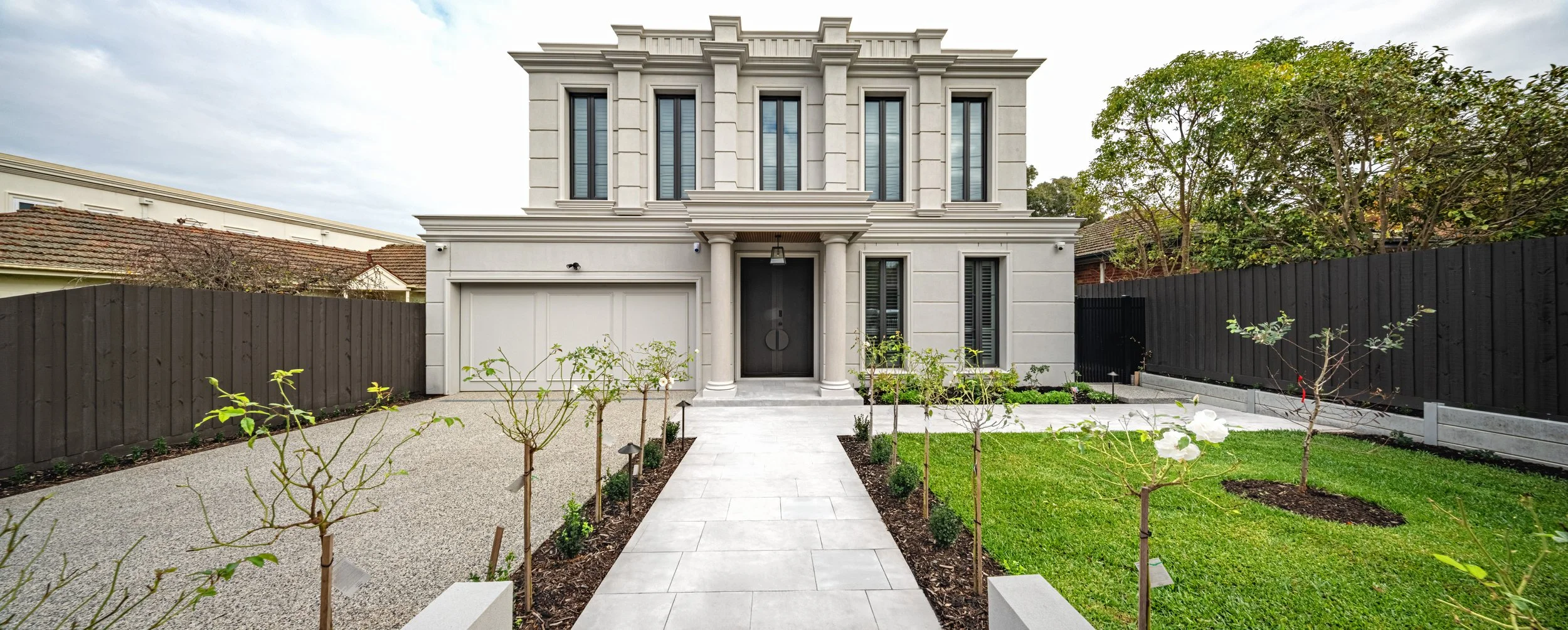 Front view of a modern two-story house with a white facade, black front door, and a small front garden with young trees and landscaped lawn, surrounded by a black wooden fence.