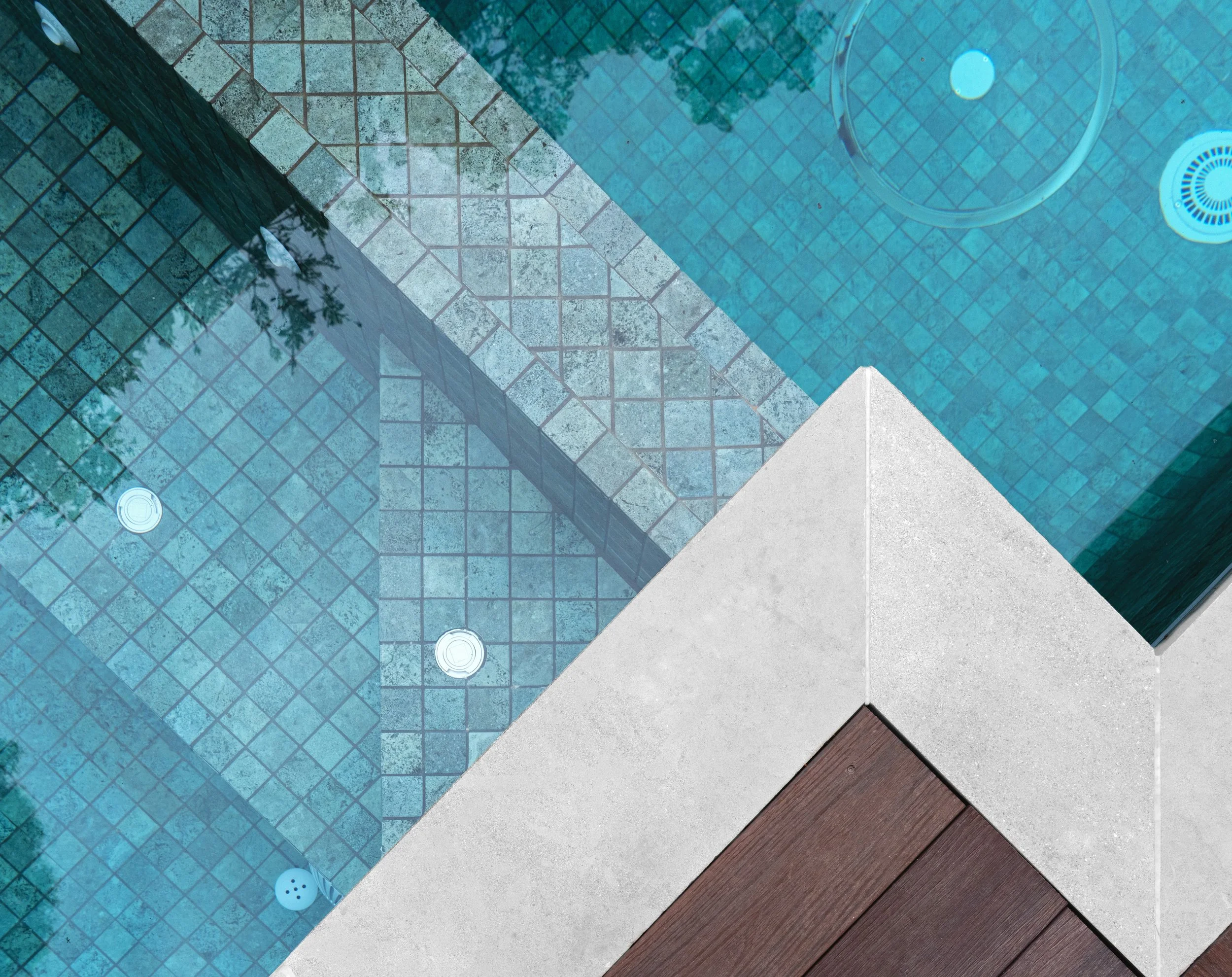 A tiled swimming pool area showing different sections of blue tile and brown wooden decking. The pool has a round drain and a white circular jet.