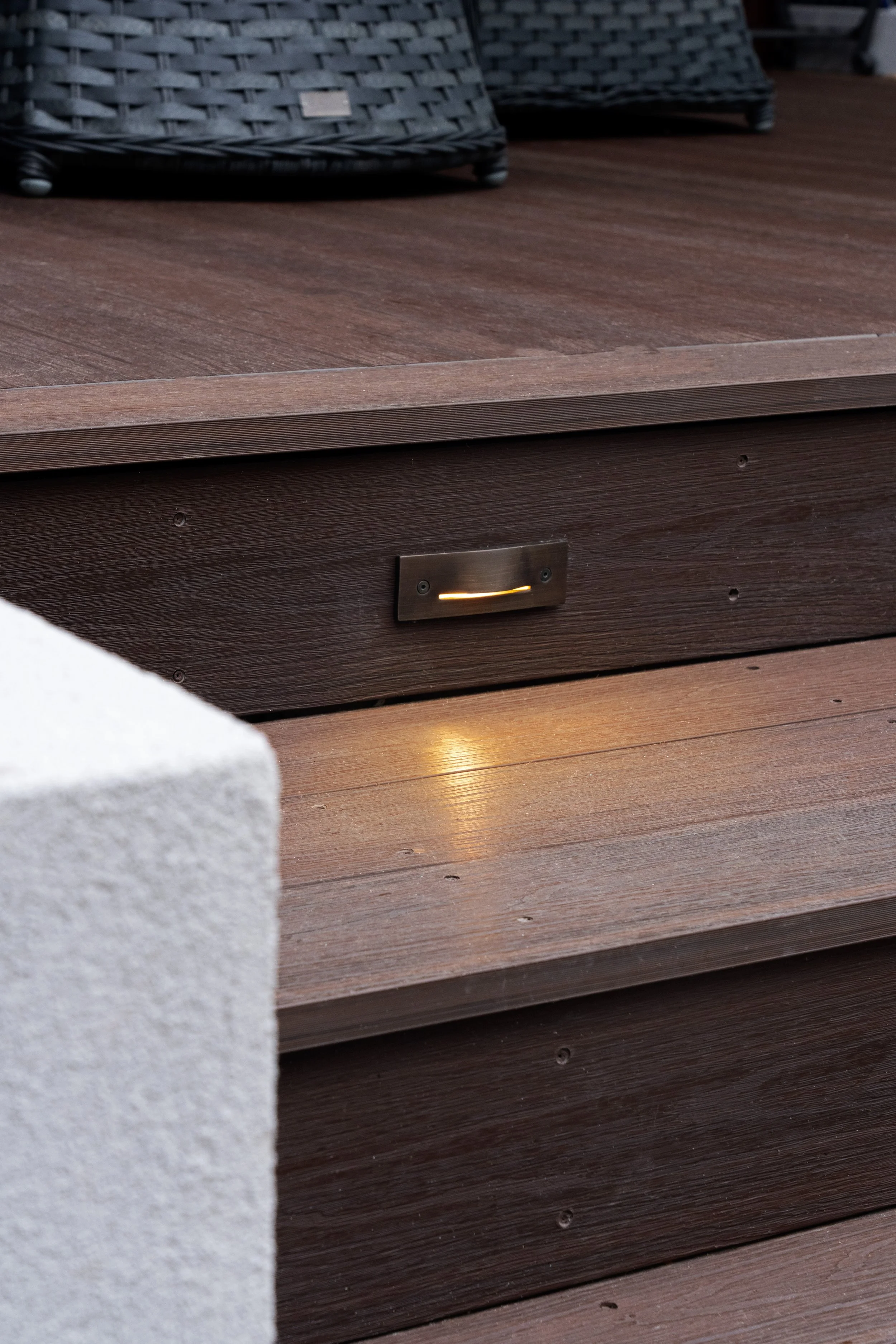 Close-up of a stepped outdoor wooden deck or patio area with wicker furniture in the background, and a small light fixture on the side of the lower level.