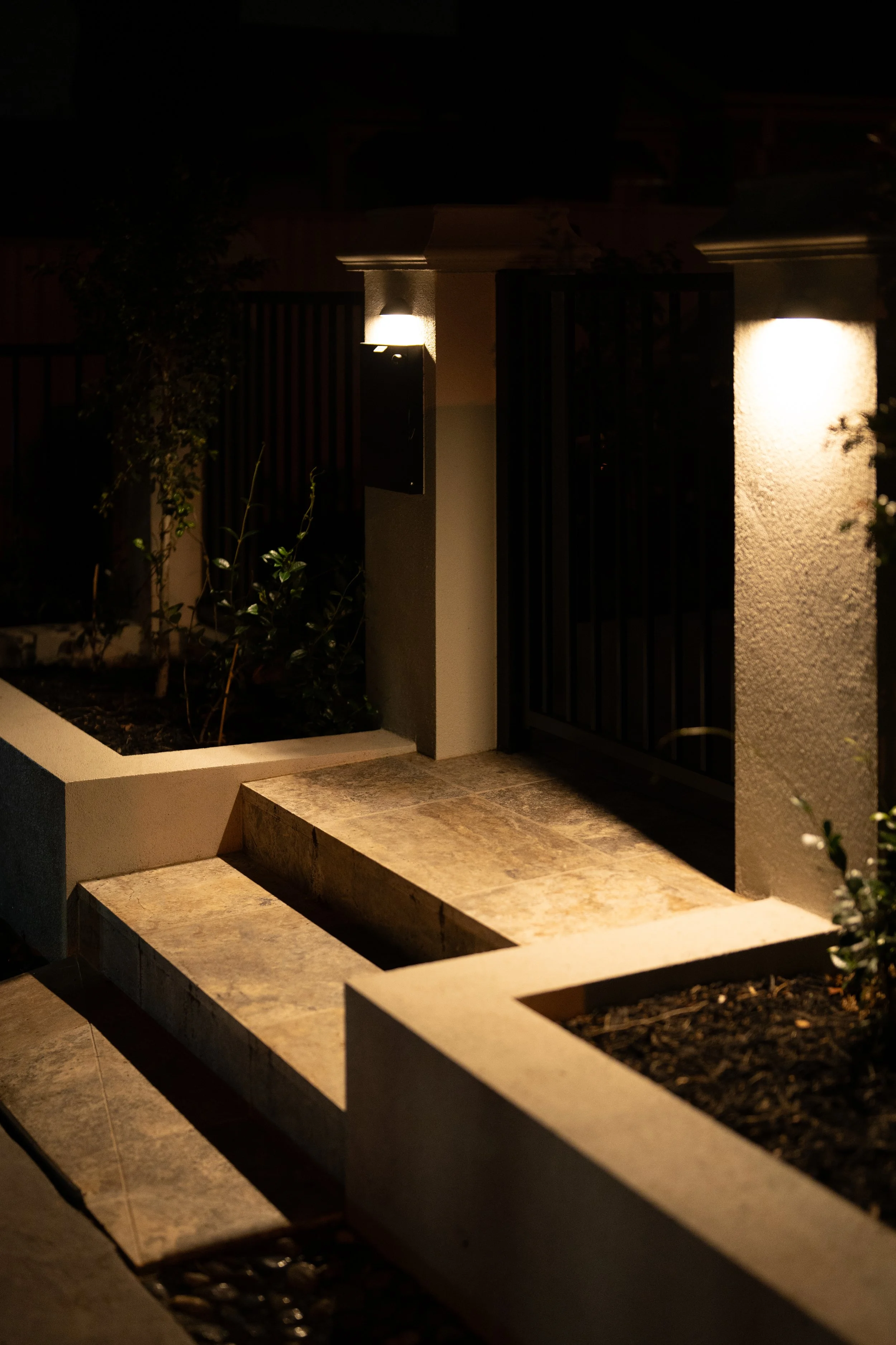 Nighttime outdoor scene of a lit entrance gate with two white pillars, a door, and a small garden with plants on the left side.