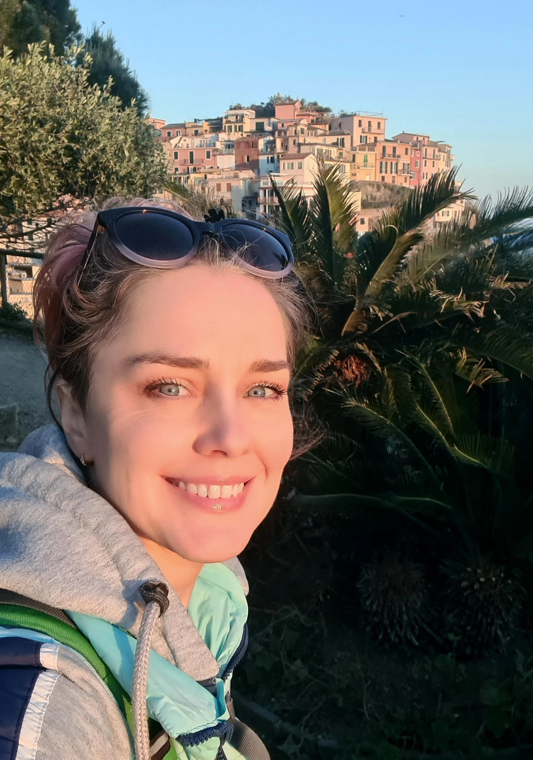 Close-up of a smiling woman with sunglasses on her head, wearing a light blue jacket and carrying a backpack, outdoors near palm trees with a colorful hillside town in the background during daytime.