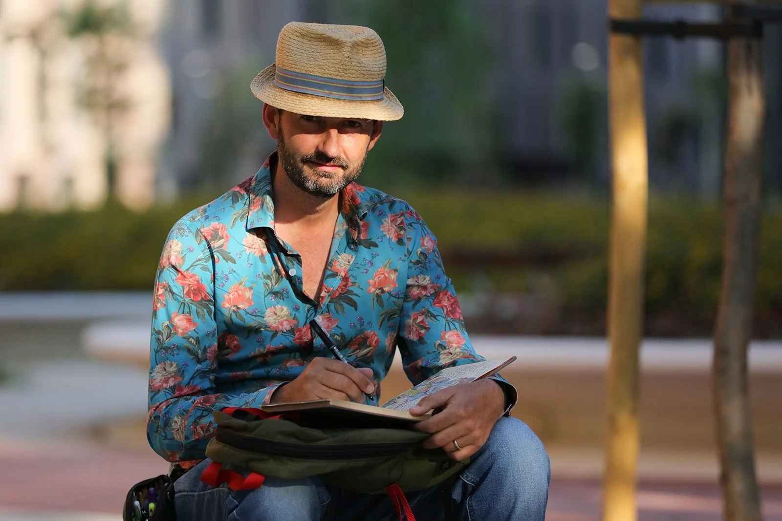 A man sitting outdoors, wearing a straw hat and a colorful floral shirt, writes in a notebook.