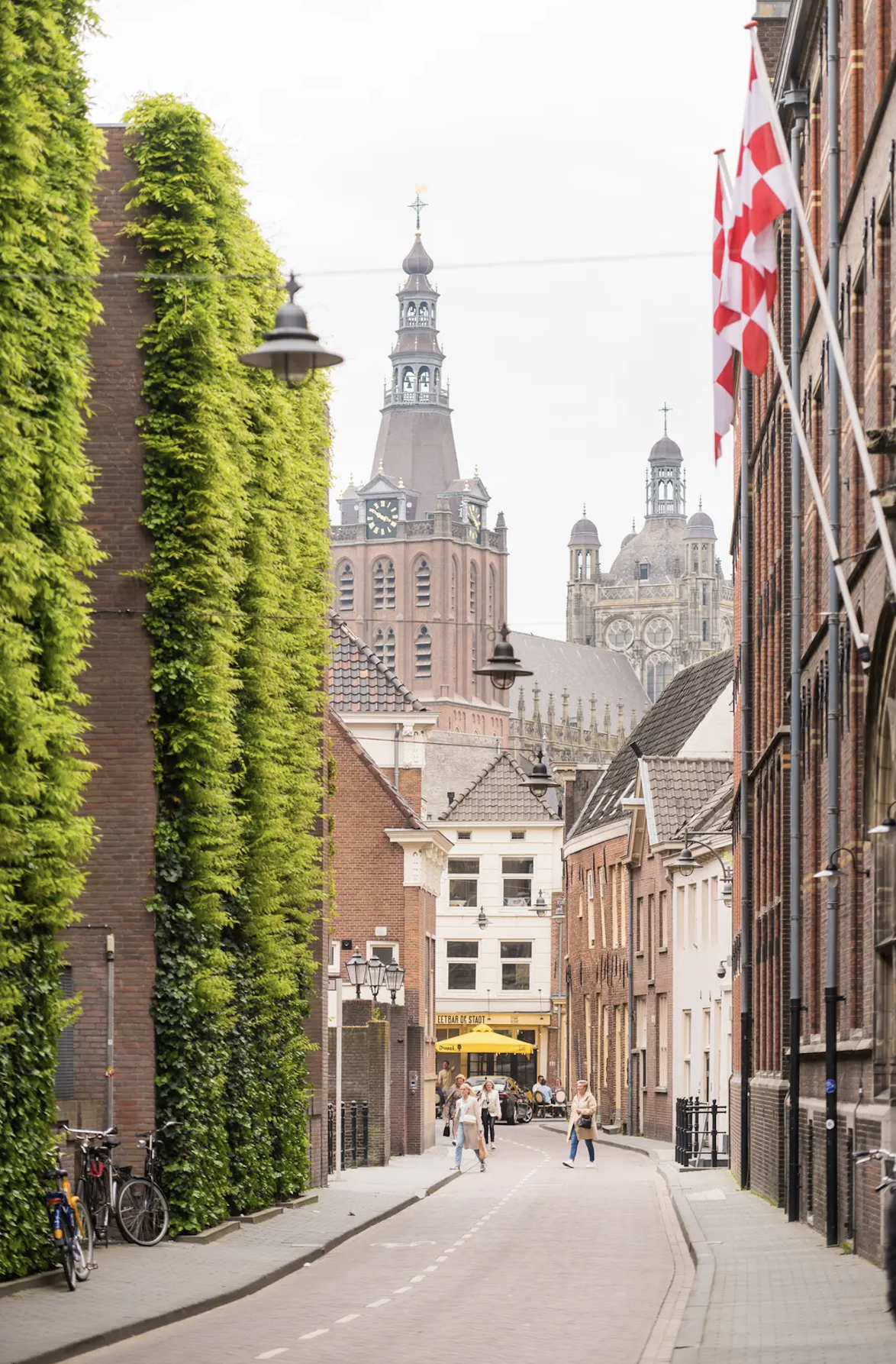 A street scene with historic buildings, a church with a tall steeple in the background, and people walking on the sidewalk. There are bicycles parked on the left and flags on the right. The street is narrow and lined with brick and white buildings.