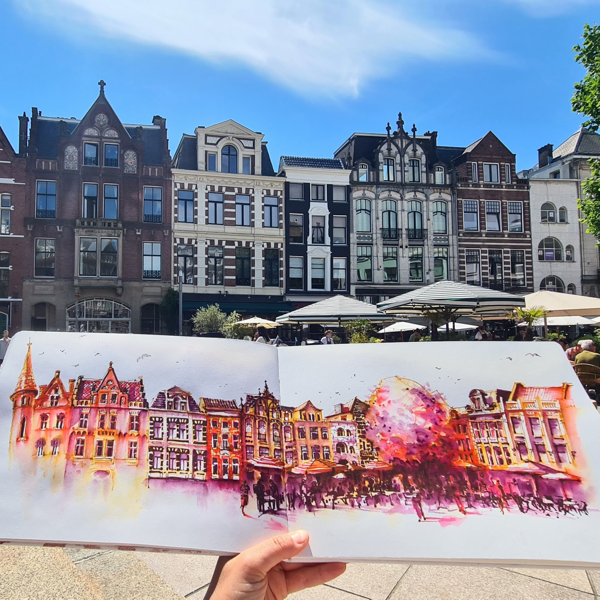 Colorful watercolor painting of European-style buildings with pointed roofs and ornate facades, held by a person outdoors with umbrellas and a clear blue sky in the background.