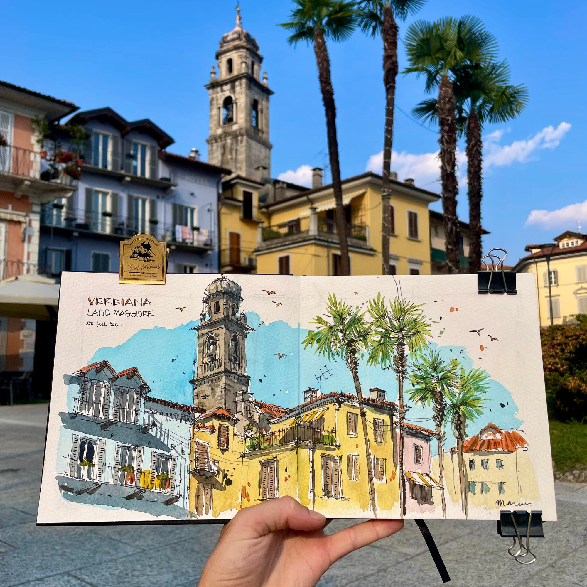 A person holding a colorful watercolor painting of a church with a tall bell tower, surrounded by palm trees, with European-style buildings in the background under a blue sky.