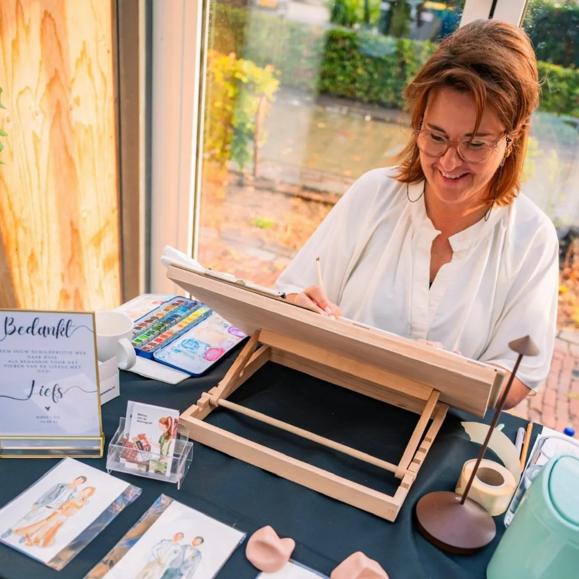 A woman with glasses and red hair smiling while painting watercolor pictures of children. Her workspace includes a watercolor palette, painted artwork, and a wooden display stand, set outdoors near autumn foliage.