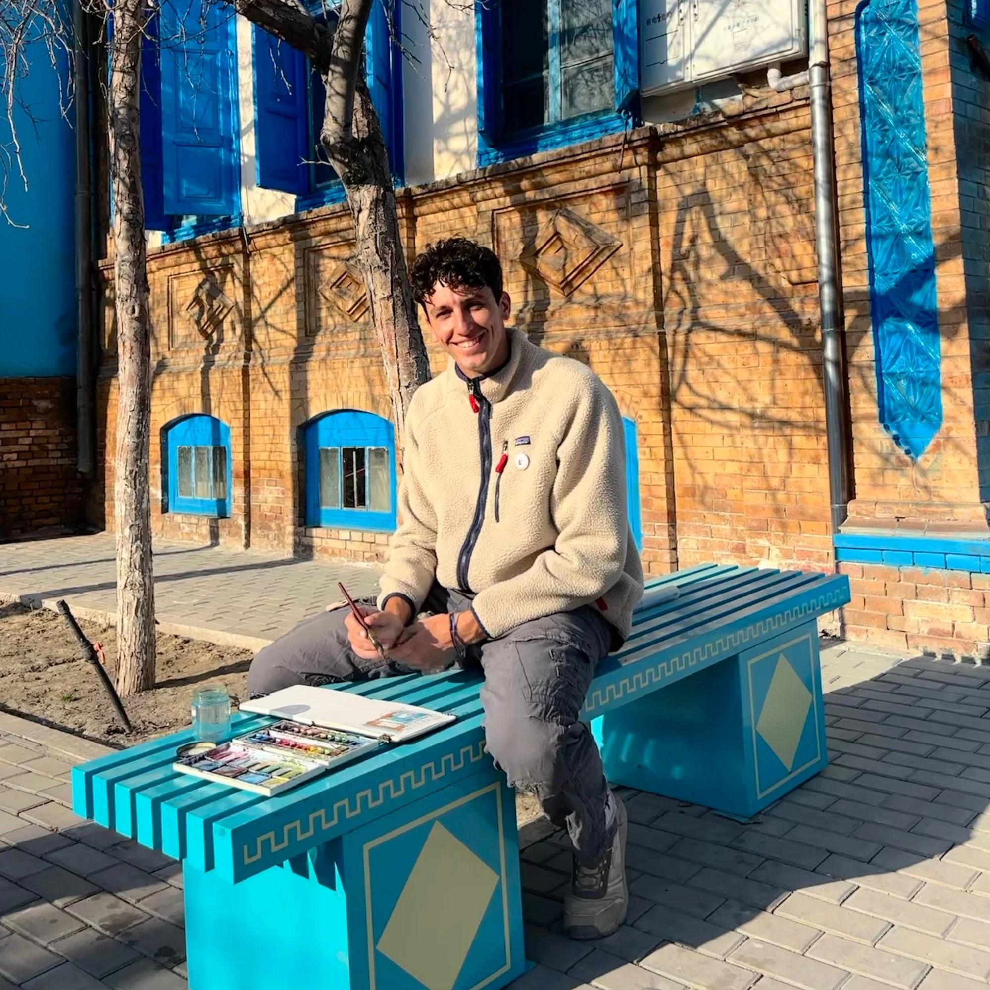 A young man with curly dark hair sitting on a blue and yellow patterned bench outdoorsn front of him is an open art set with paints and brushes. Behind him, there is a brick building with blue window frames and shutters, a mailbox, a leafless tree, and a clear sky.