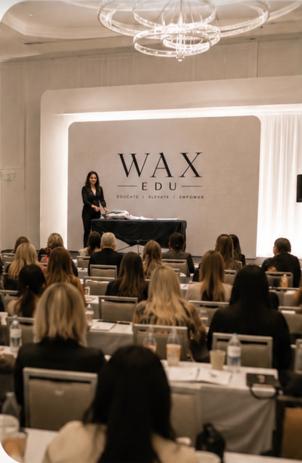 A speaker presenting on stage at a conference called WAX EDU, where the slogan is Educate, Elevate, Empower. The audience is seated and listening in a large room with a high ceiling and modern lighting fixture.