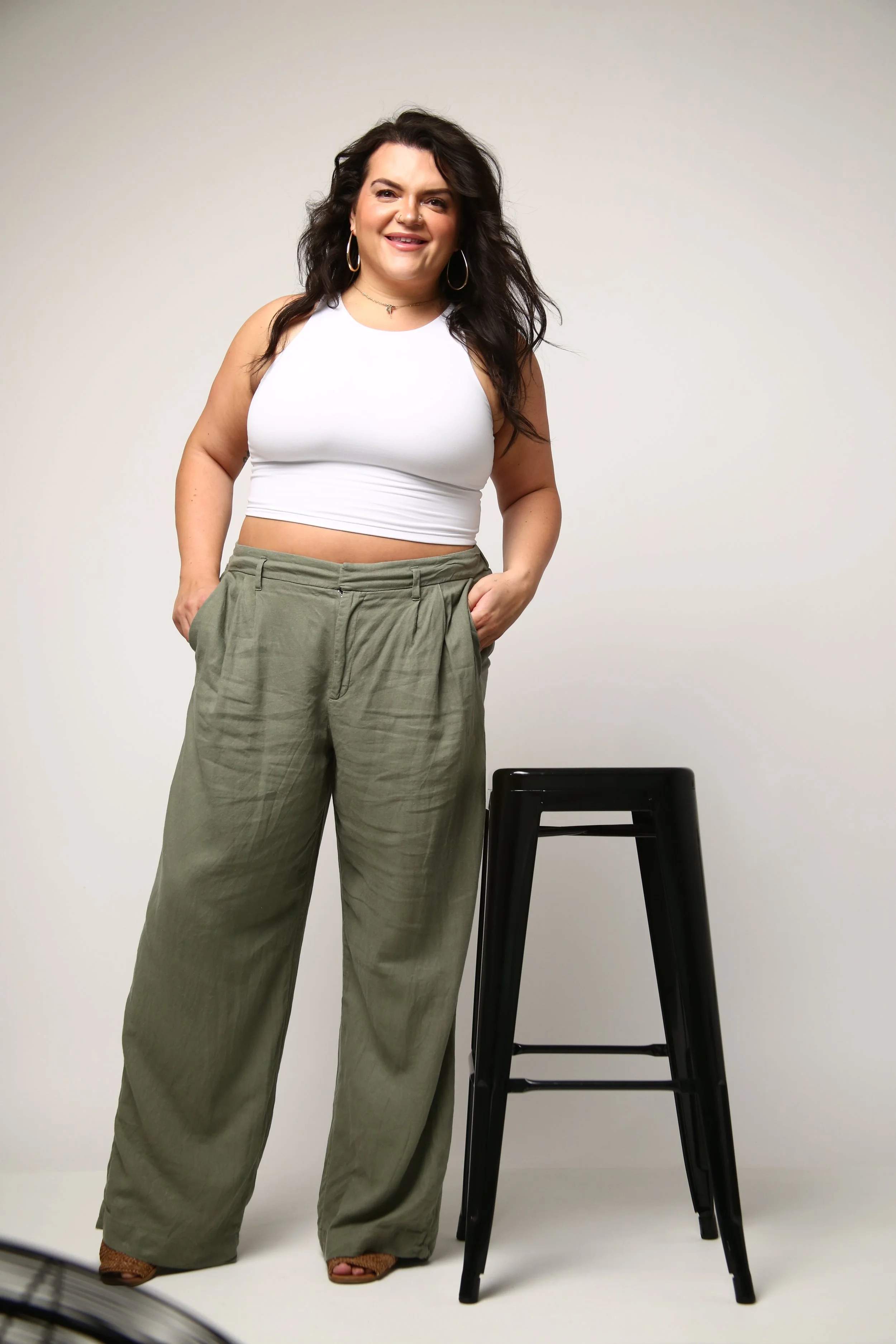A woman with long, dark, curly hair, smiling and wearing hoop earrings, a choker necklace, a sleeveless white crop top, wide-legged green pants, and brown sandals standing next to a black stool against a plain light-colored background.