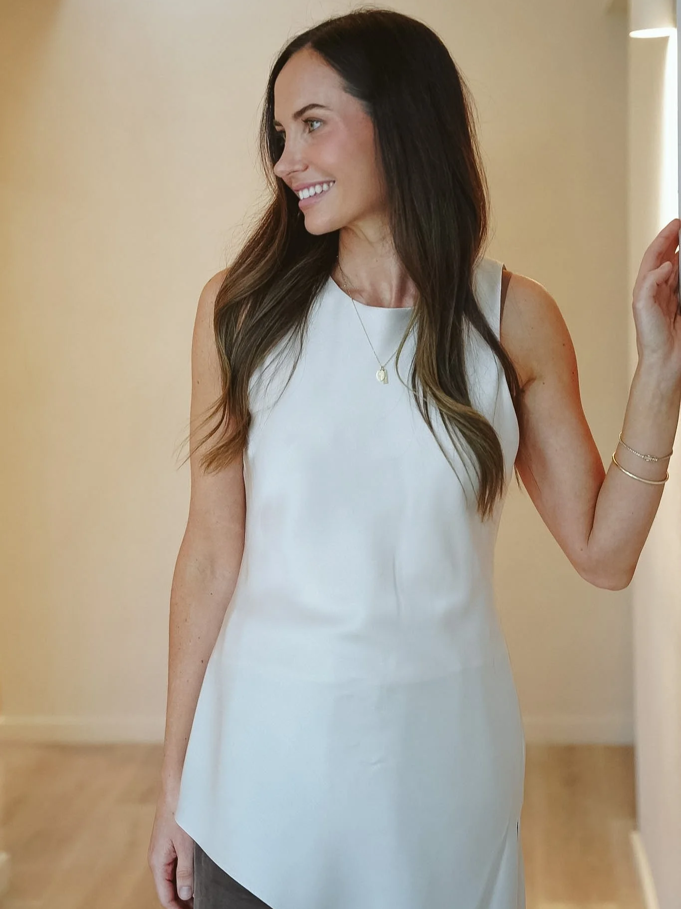 Jessica Stacey, founder of Forme Cosmetic Science,A woman with long dark hair smiling, wearing a white sleeveless top, a gold necklace with a small pendant, and a gold bracelet, standing indoors against a beige wall with wooden flooring.